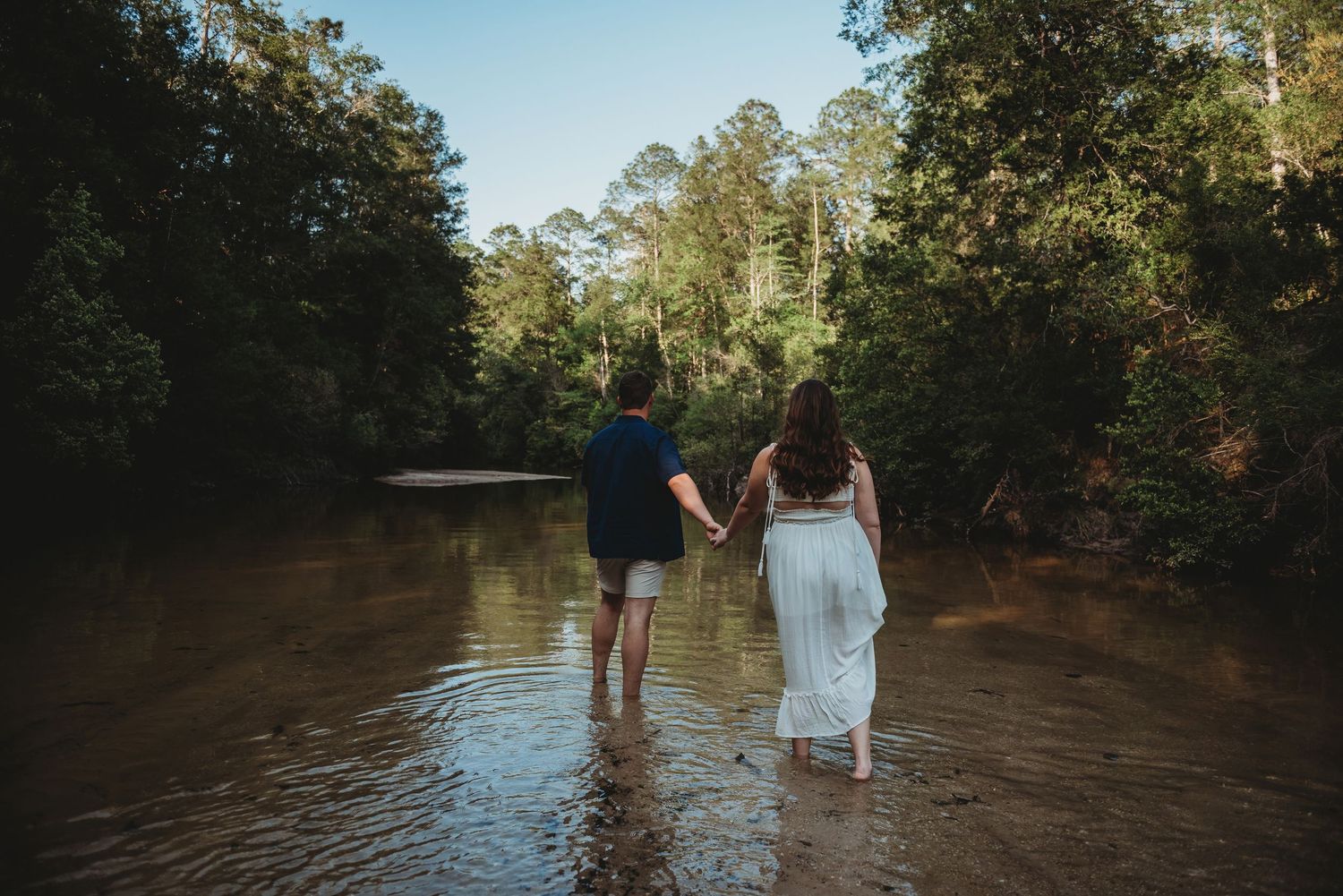 Couple holding hands walking through a shallow forest river with tall green trees.