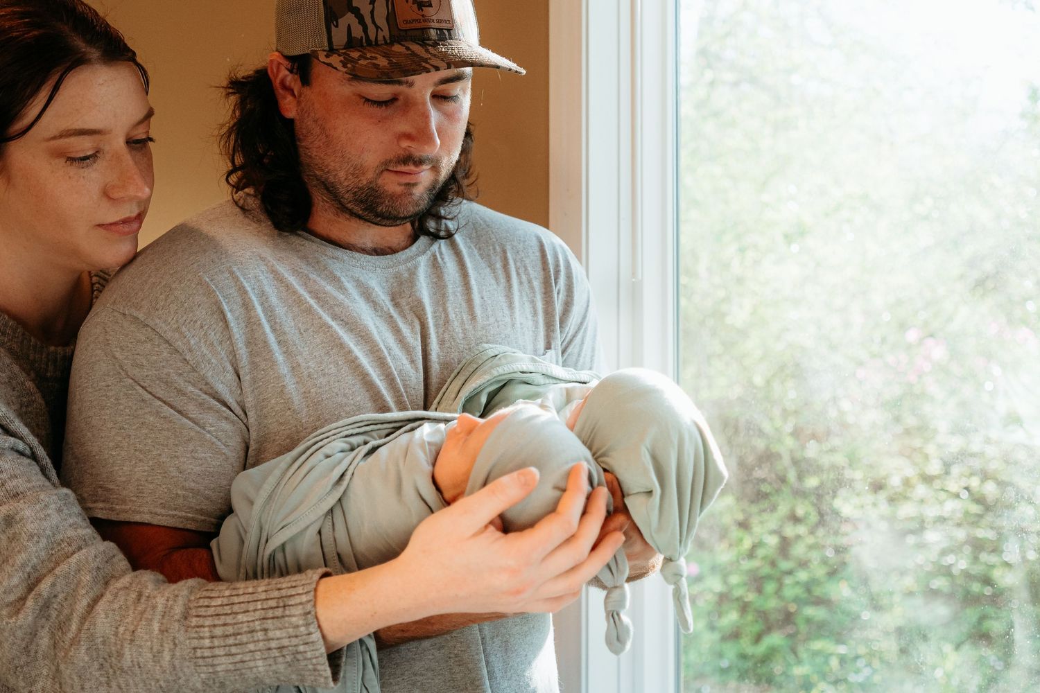 New parents tenderly hold their swaddled newborn baby together by a bright window.
