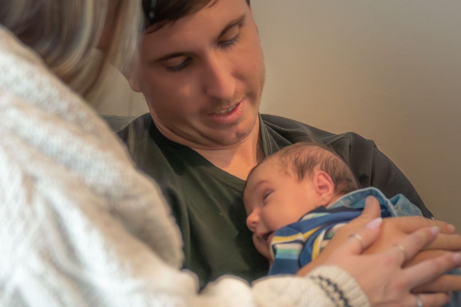 Smiling father cradling sleeping newborn as mother looks on lovingly in soft light.