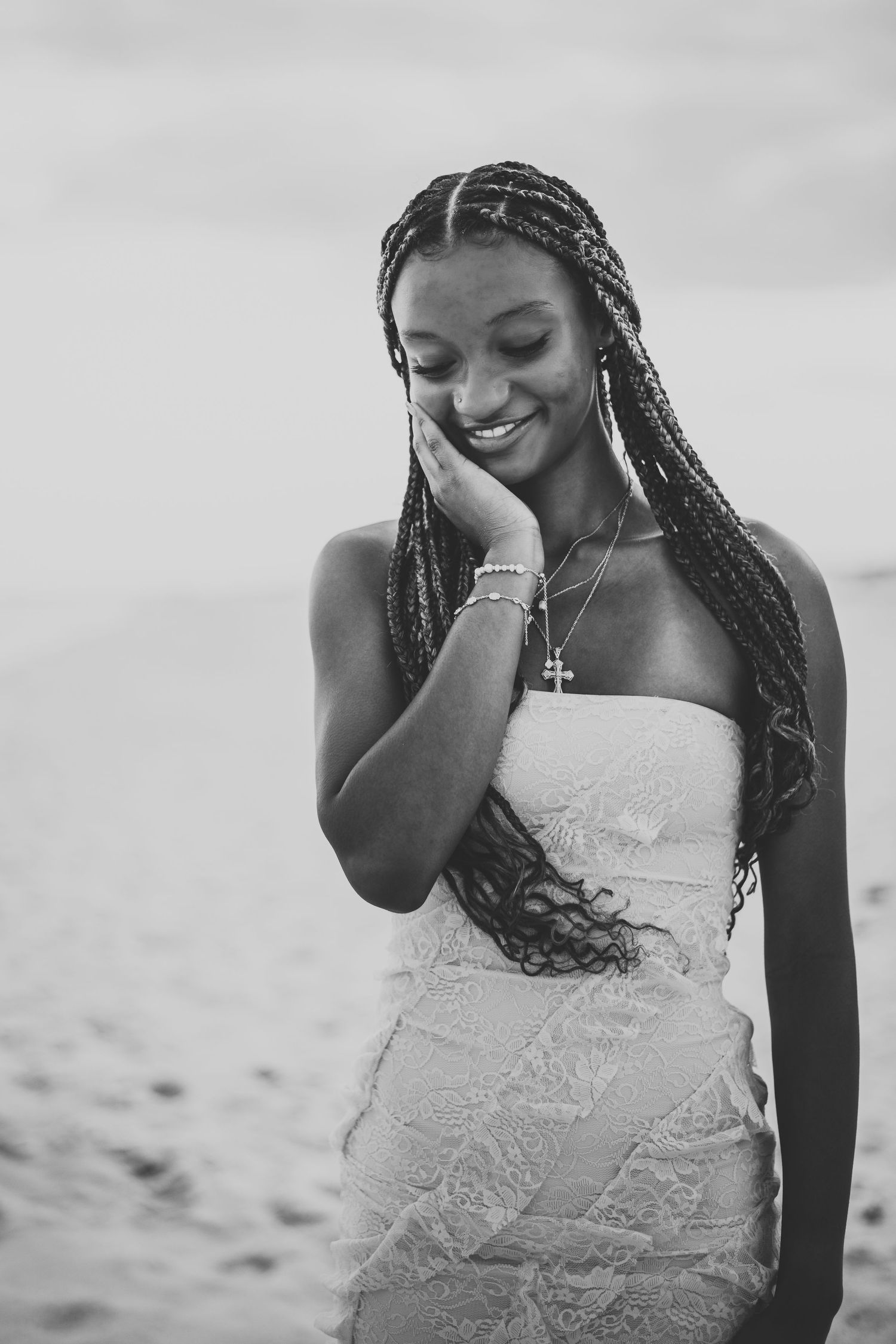 Black and white portrait of a smiling young woman with long braids in a strapless dress on a beach.