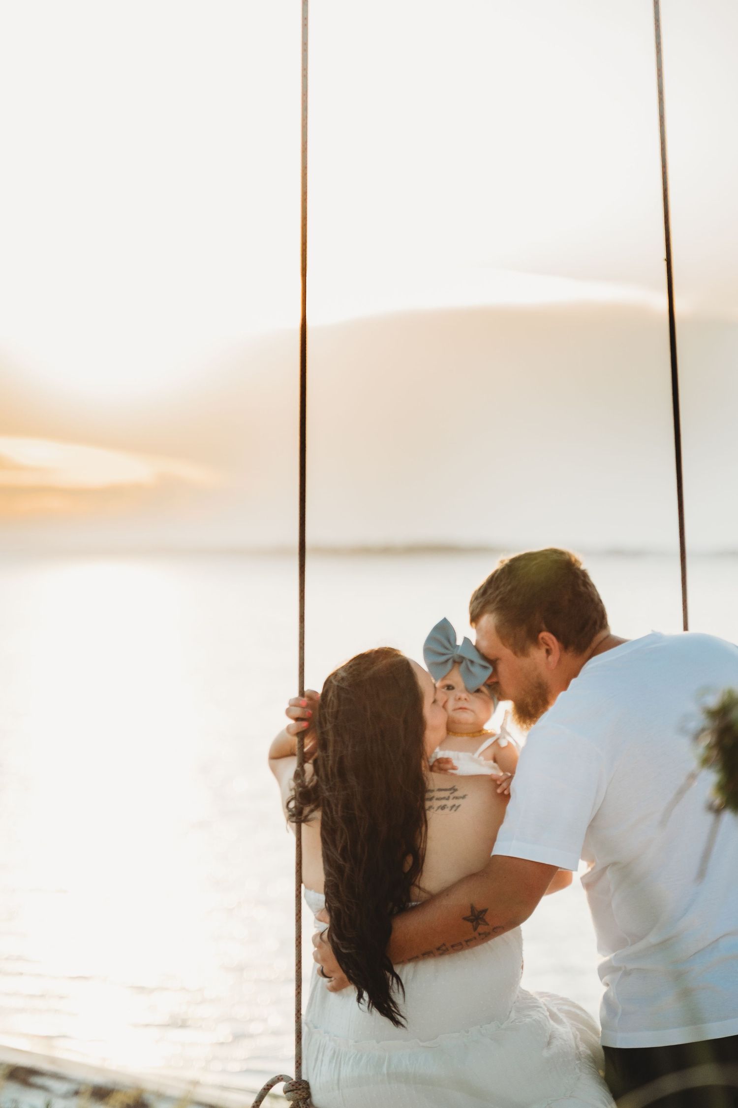 A couple kisses their baby girl on a swing by the water at golden hour.