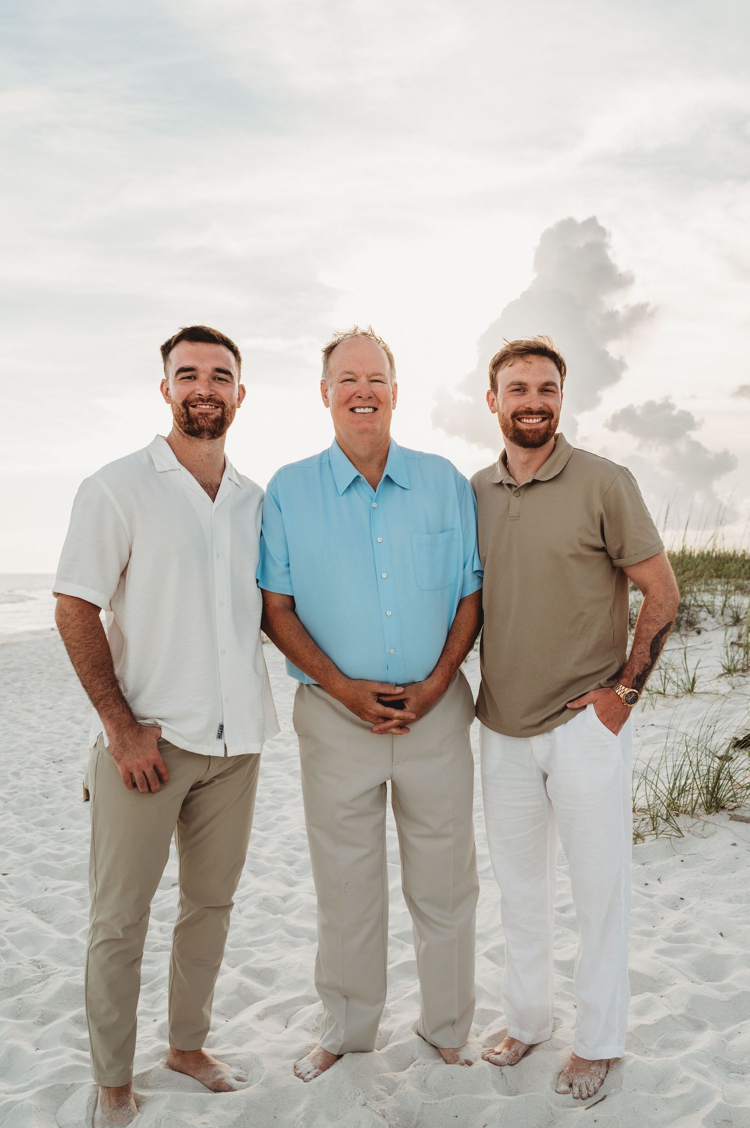 Three men, father and two sons, smiling together on a white sandy beach.