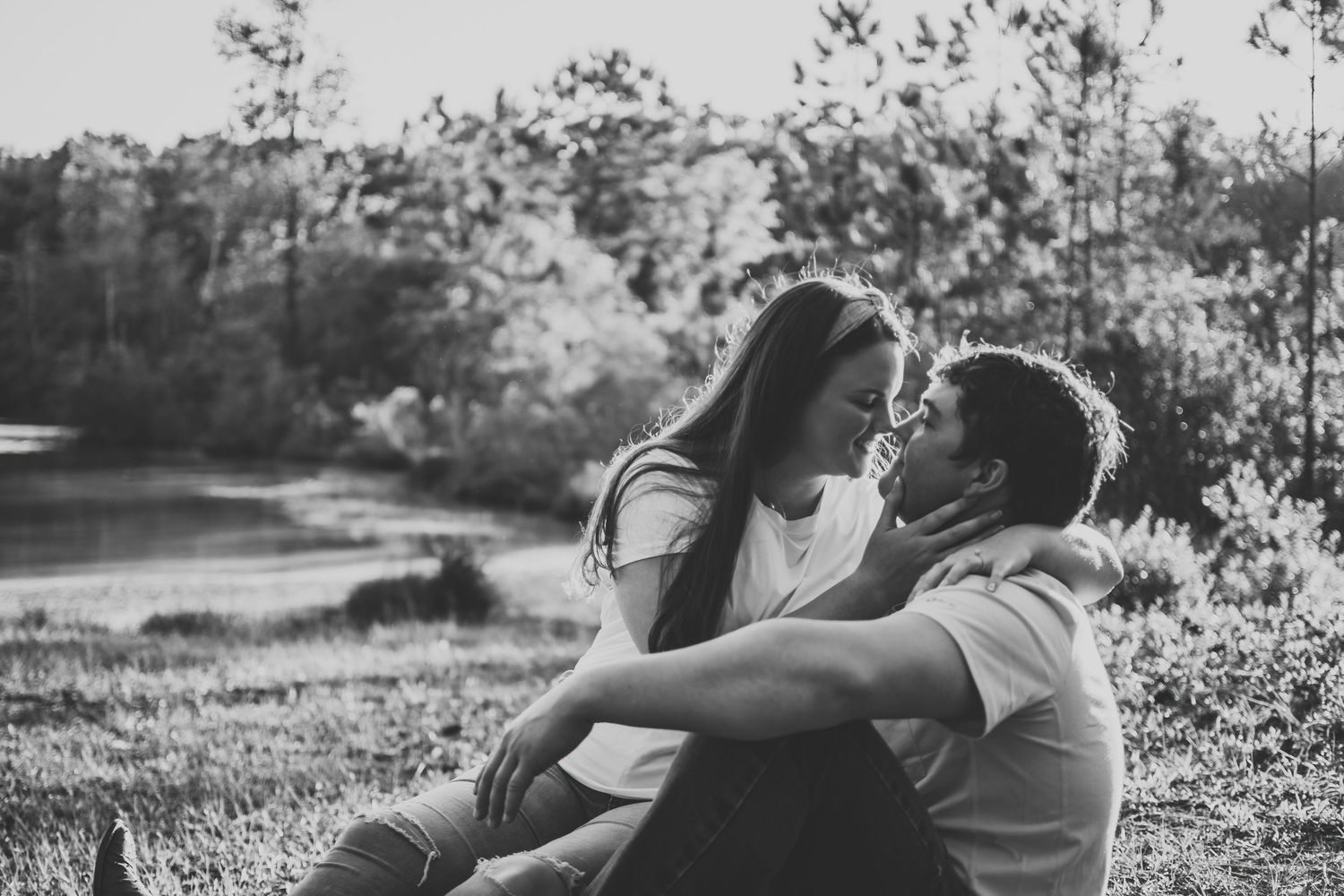 Black and white photo of a romantic couple sharing an intimate moment by a serene lake outdoors.