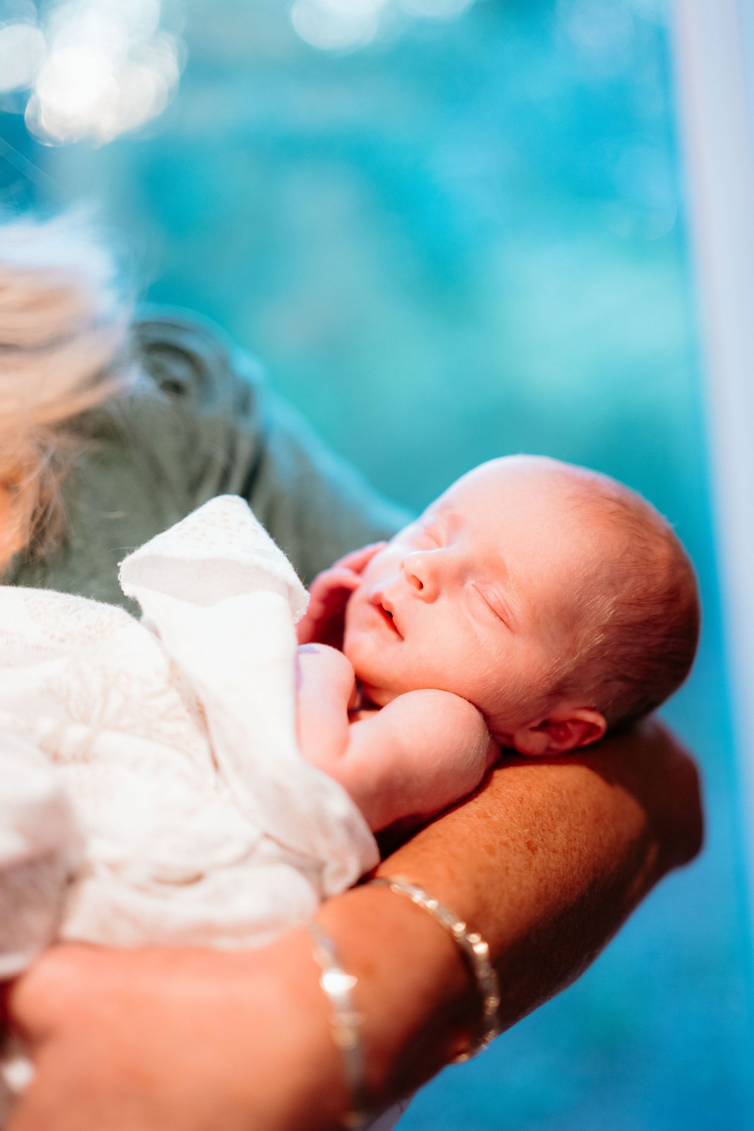 Grandmother holding sleeping newborn baby wrapped in white blanket near bright window.