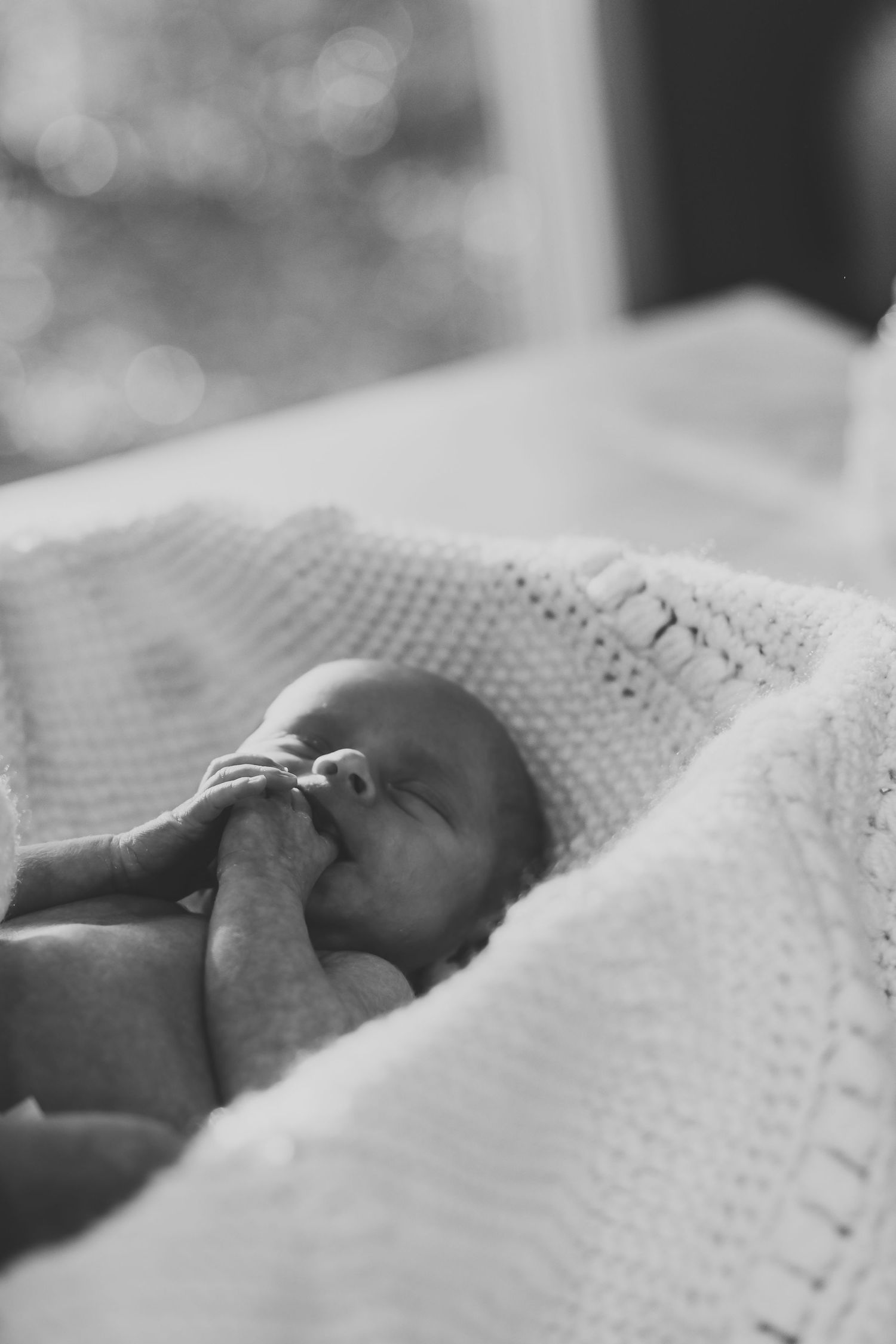 Black and white photo of a sleeping newborn baby wrapped in a knit blanket with tiny hands near face.