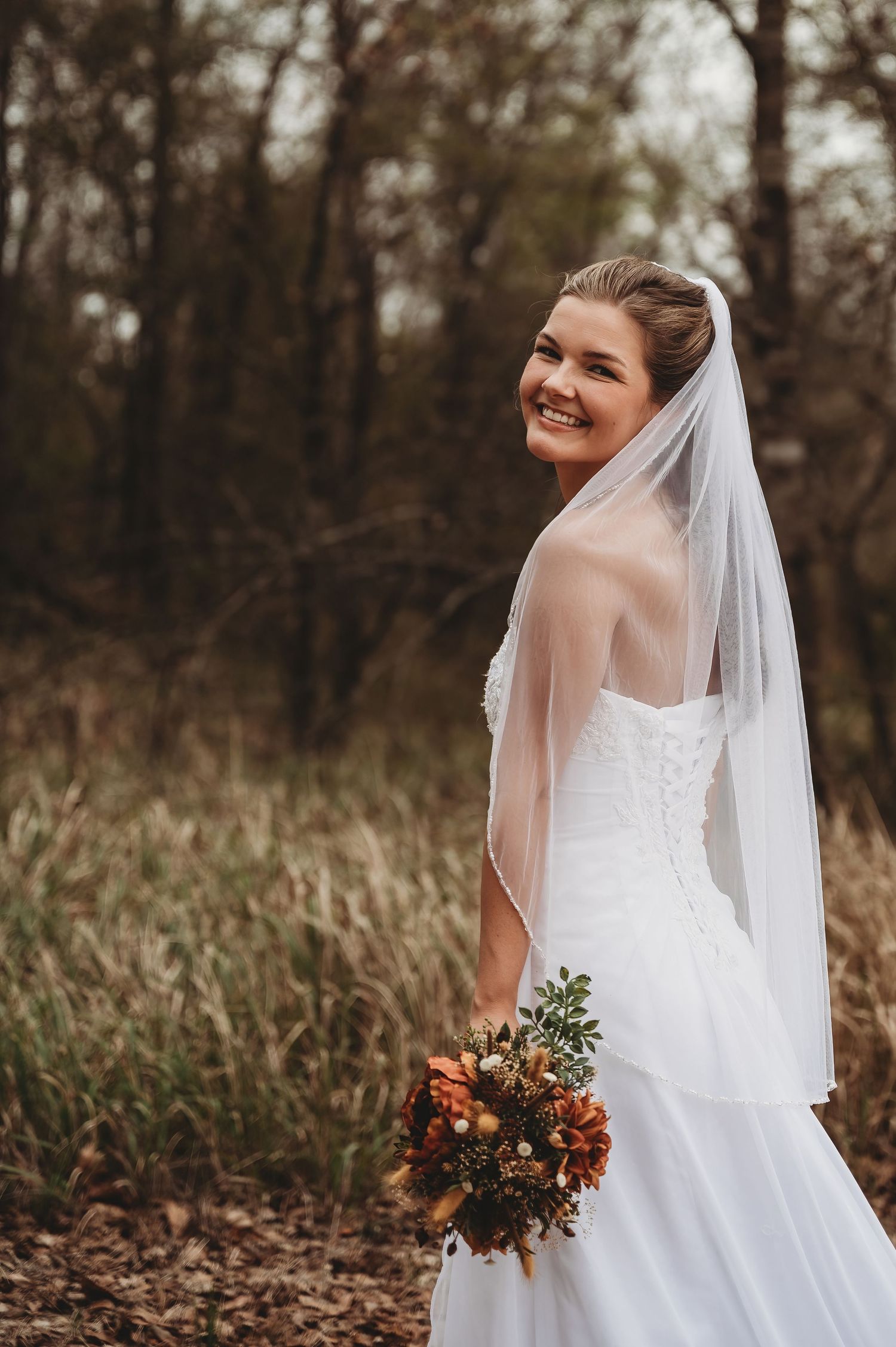 Smiling bride in white strapless gown and veil holds autumn bouquet outdoors among fall foliage.