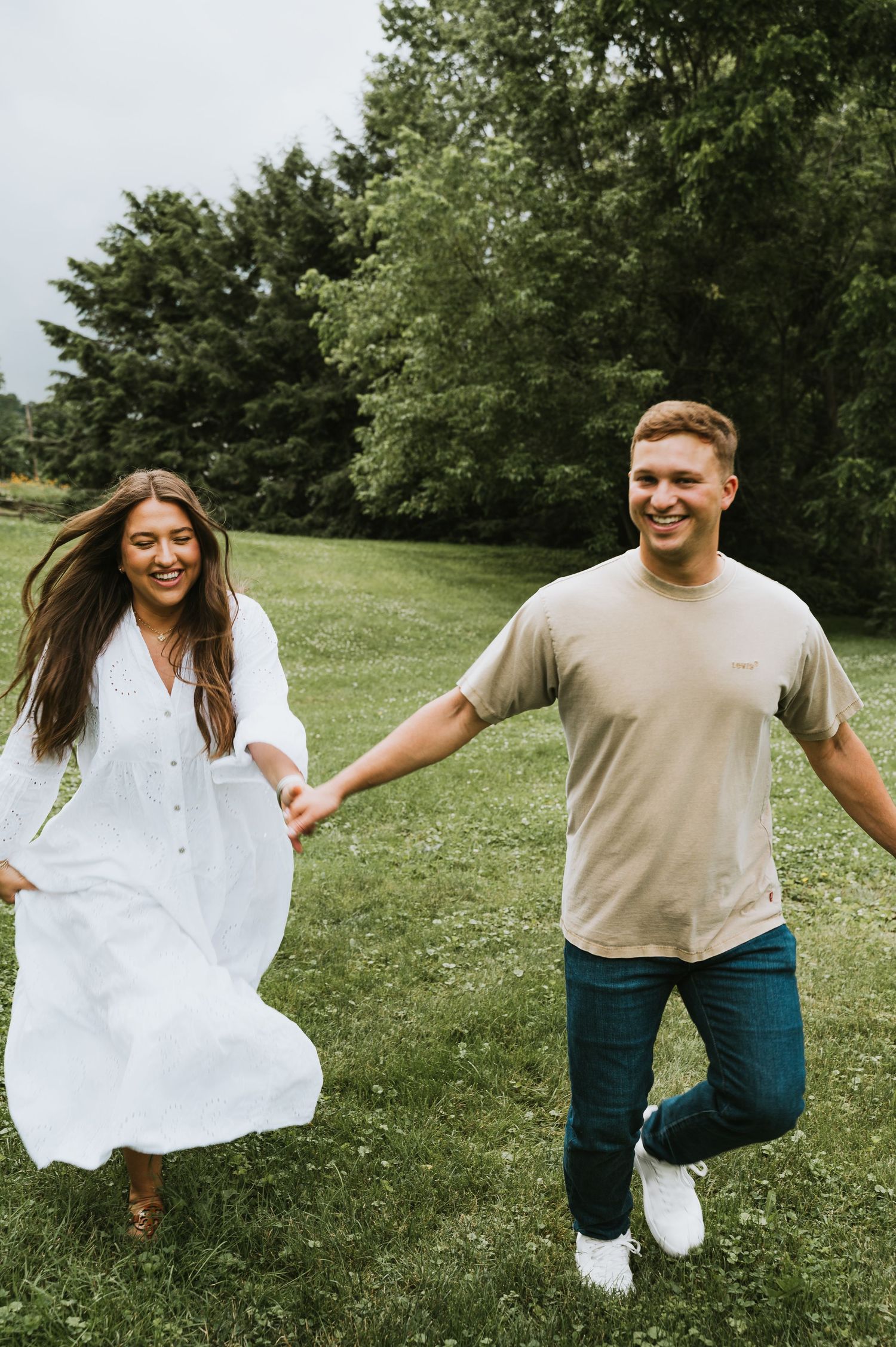Happy couple holding hands and running through a lush green park, woman in white dress, man in beige shirt.