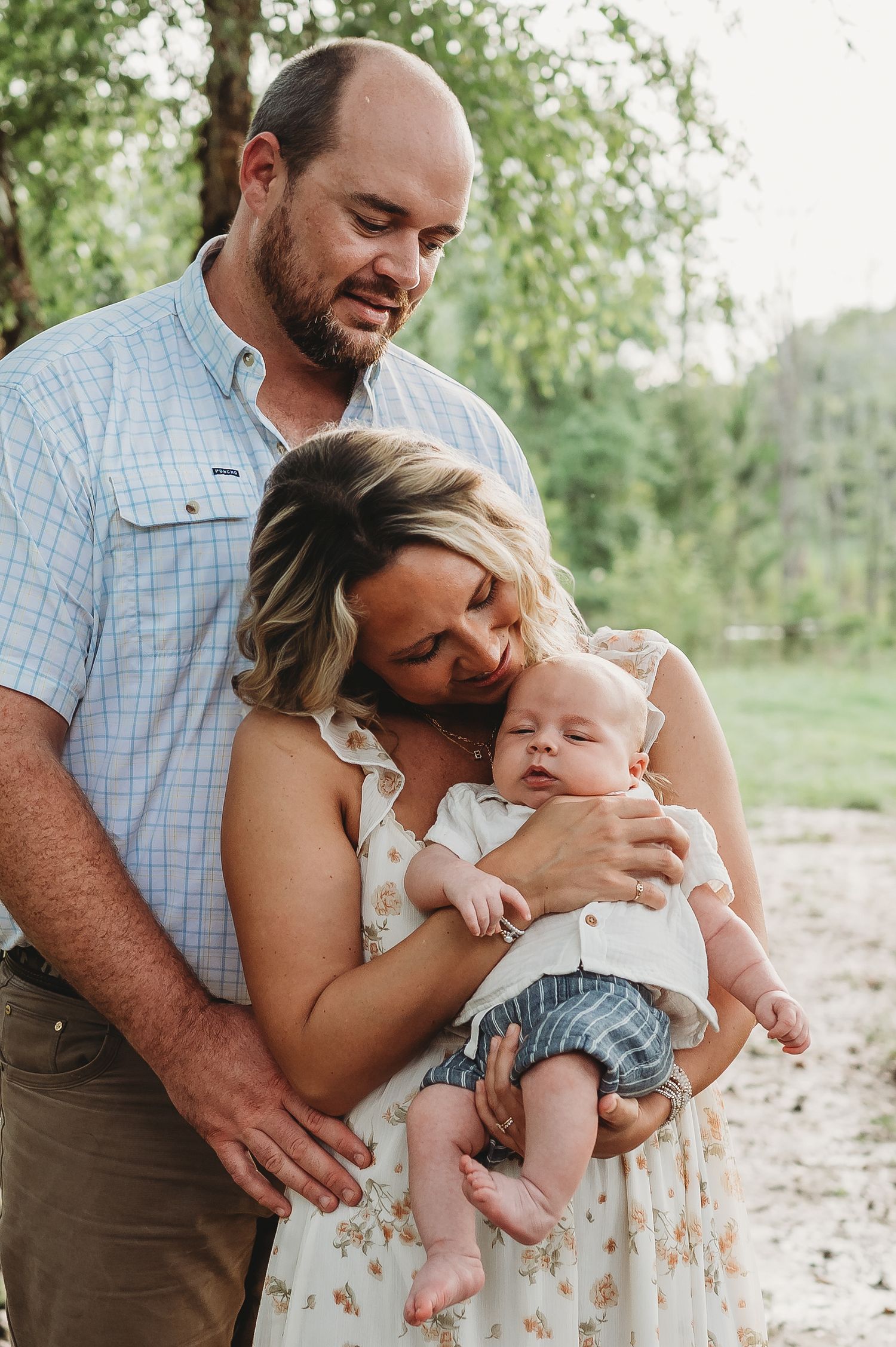 A couple tenderly holds their newborn baby outdoors surrounded by lush green trees in a warm family portrait.