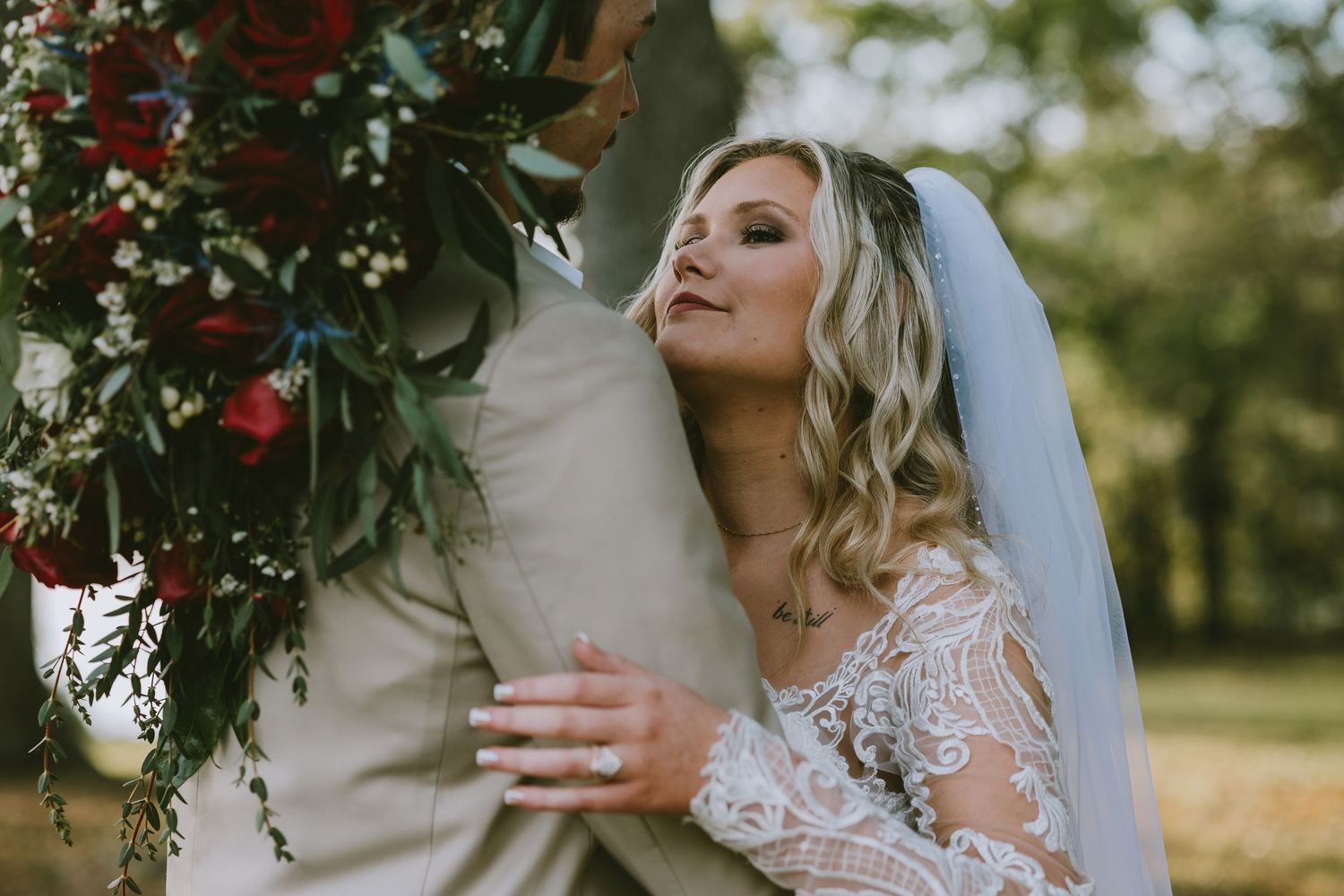 Bride in lace gown and veil embraces groom outdoors, holding red floral bouquet during romantic wedding ceremony.