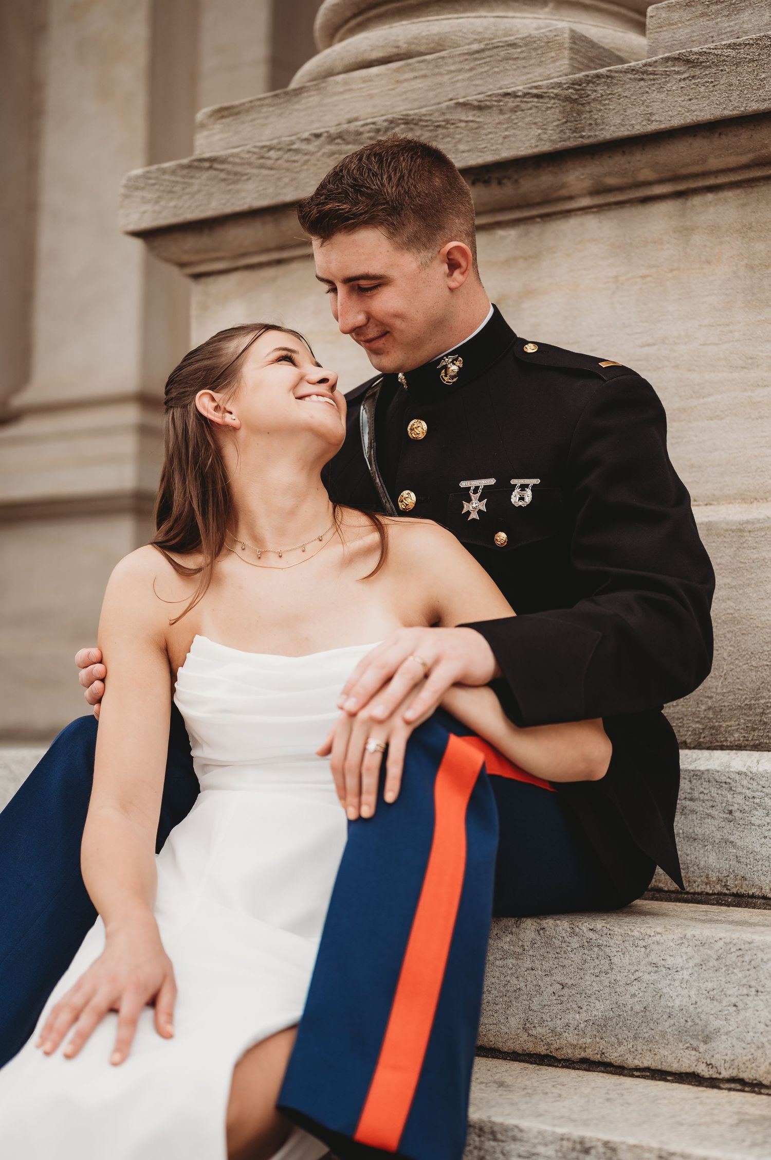 Laughing bride leans into Marine groom while sitting on marble steps in warm sunlight.