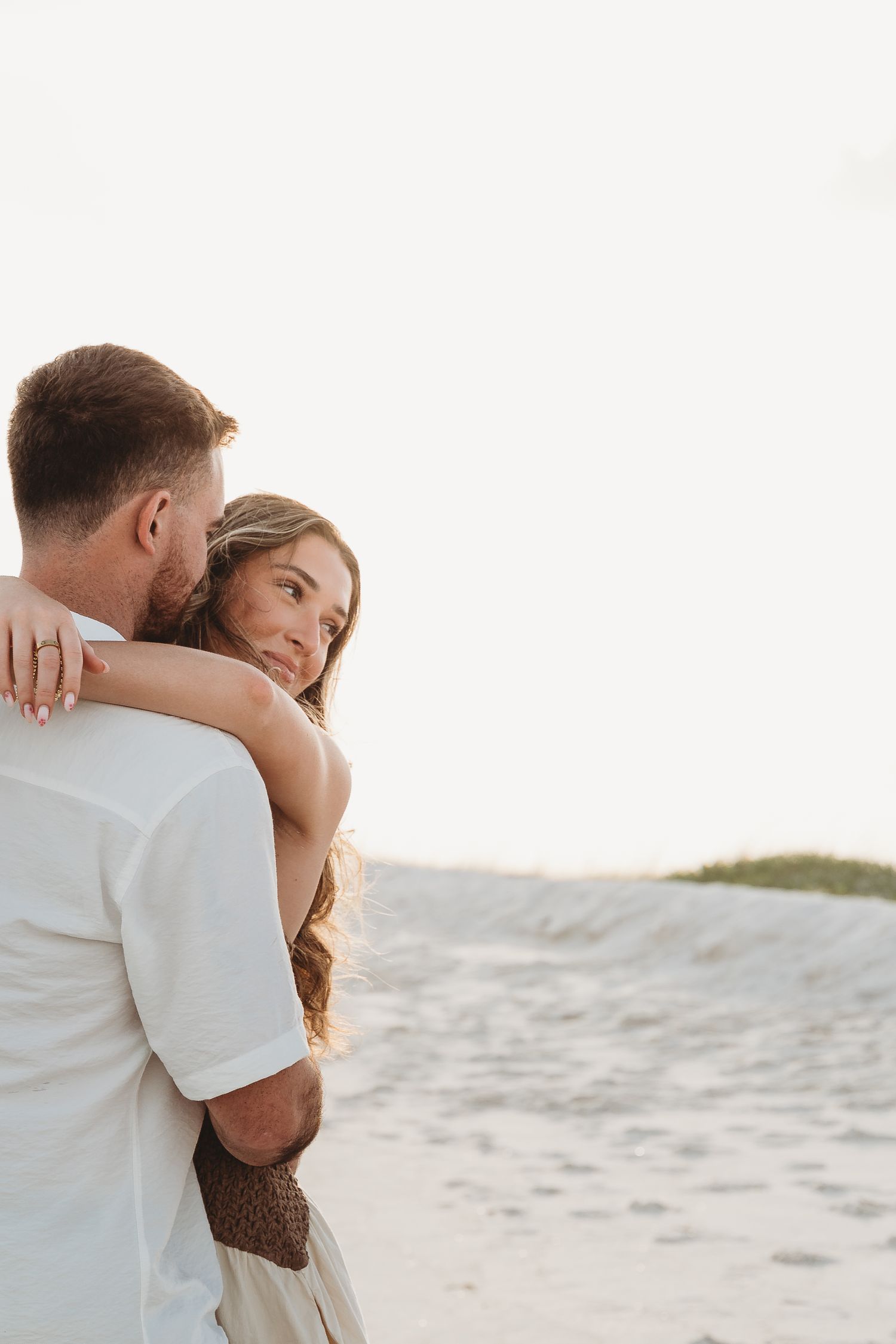 A couple shares a tender embrace on a sandy beach, woman looking up at man near sand dunes.