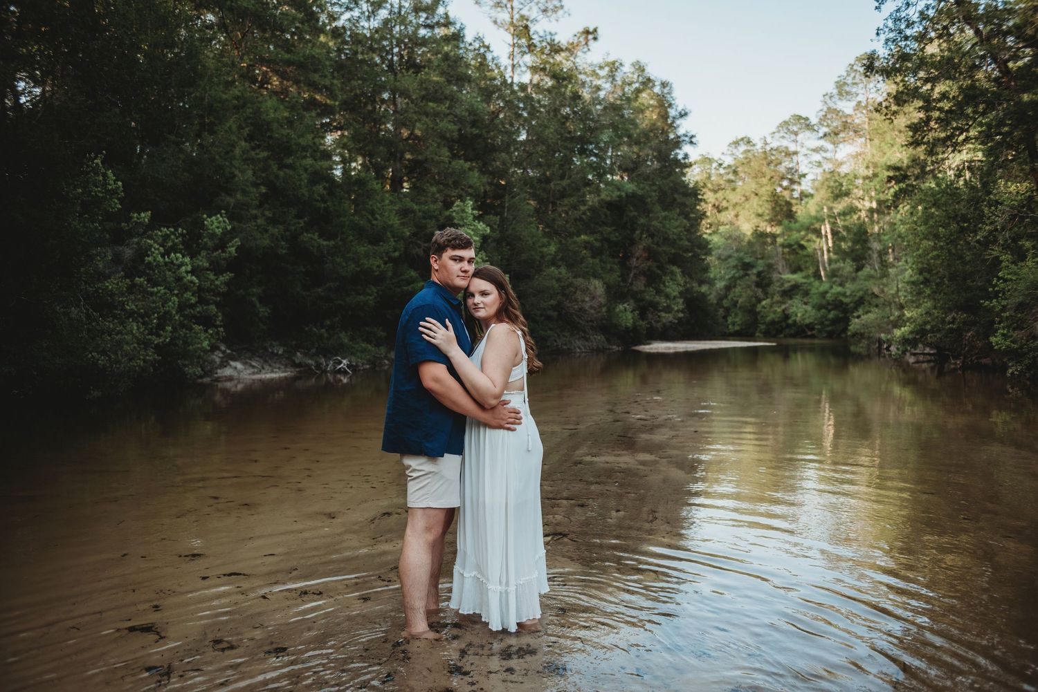 Couple standing together in shallow river water, woman in white dress hugging man in blue shirt.