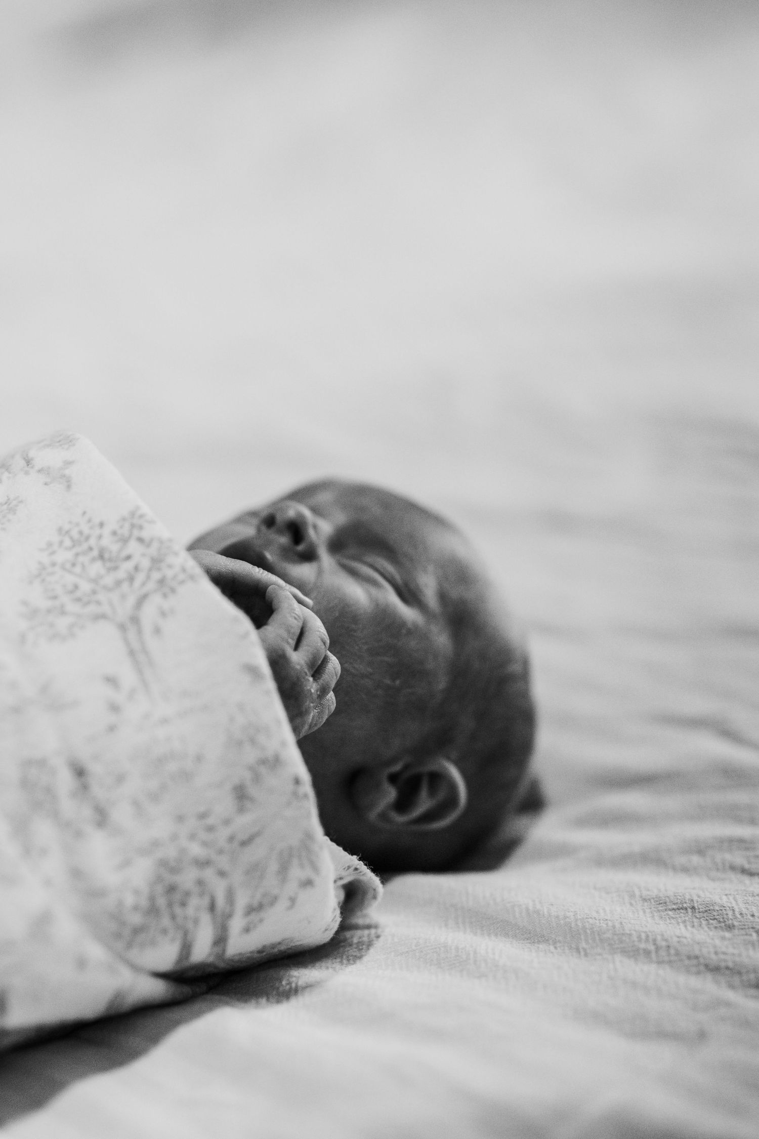Black and white close-up of a newborn baby yawning while swaddled in a patterned muslin blanket.