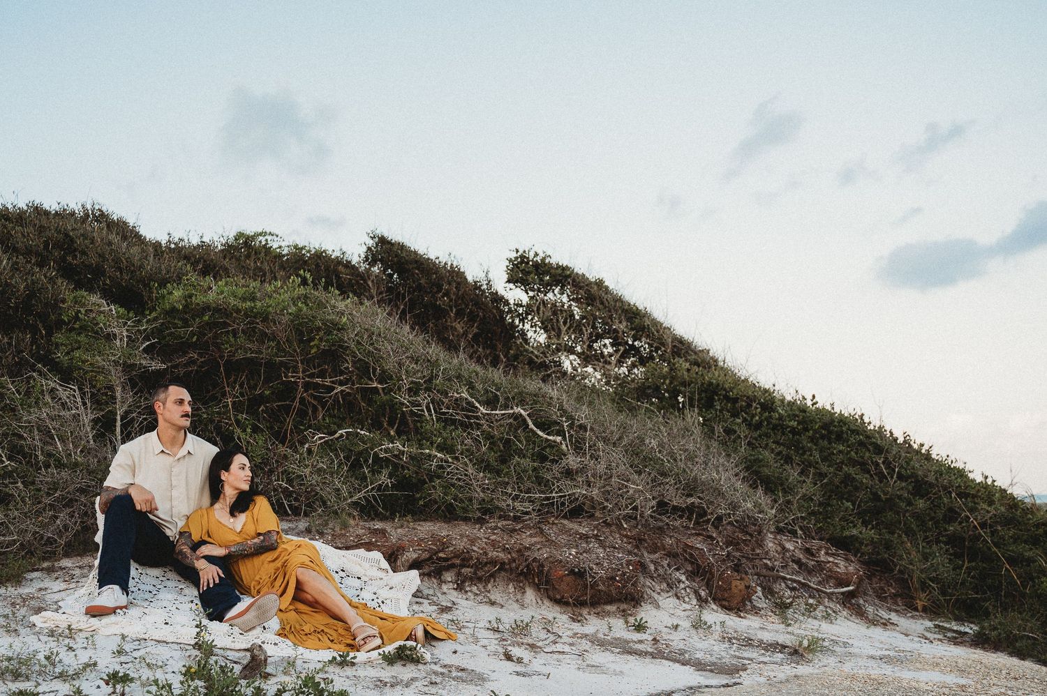 Couple sitting on white sand dunes surrounded by lush green vegetation during golden hour.