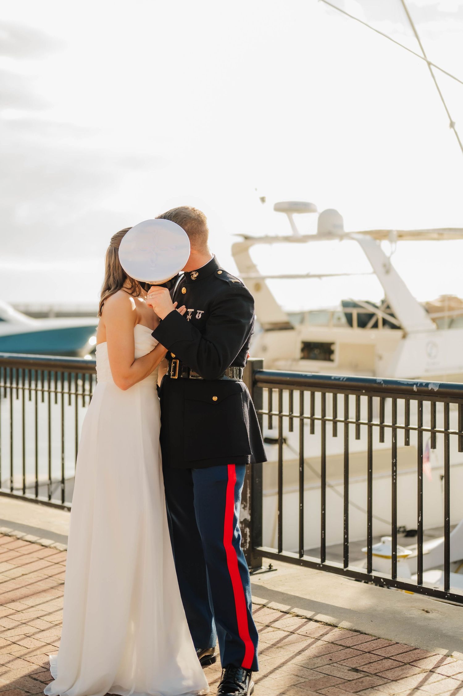 Marine in dress uniform kissing bride in white gown on waterfront dock with boats in background.