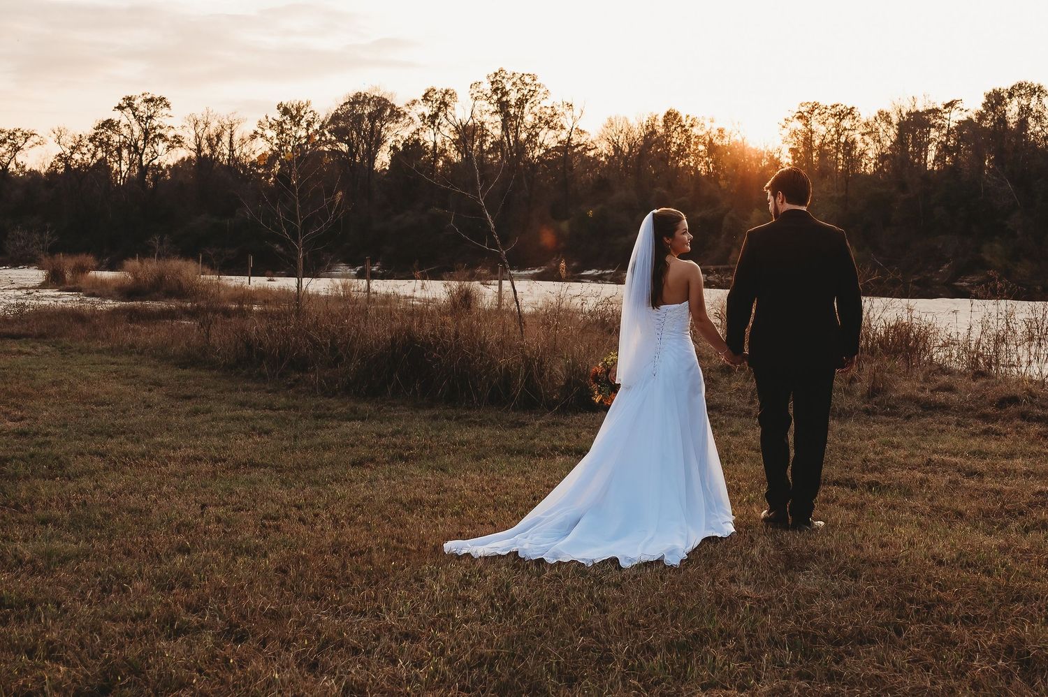 Bride and groom holding hands walking toward a scenic sunset lake surrounded by autumn trees in an open field.