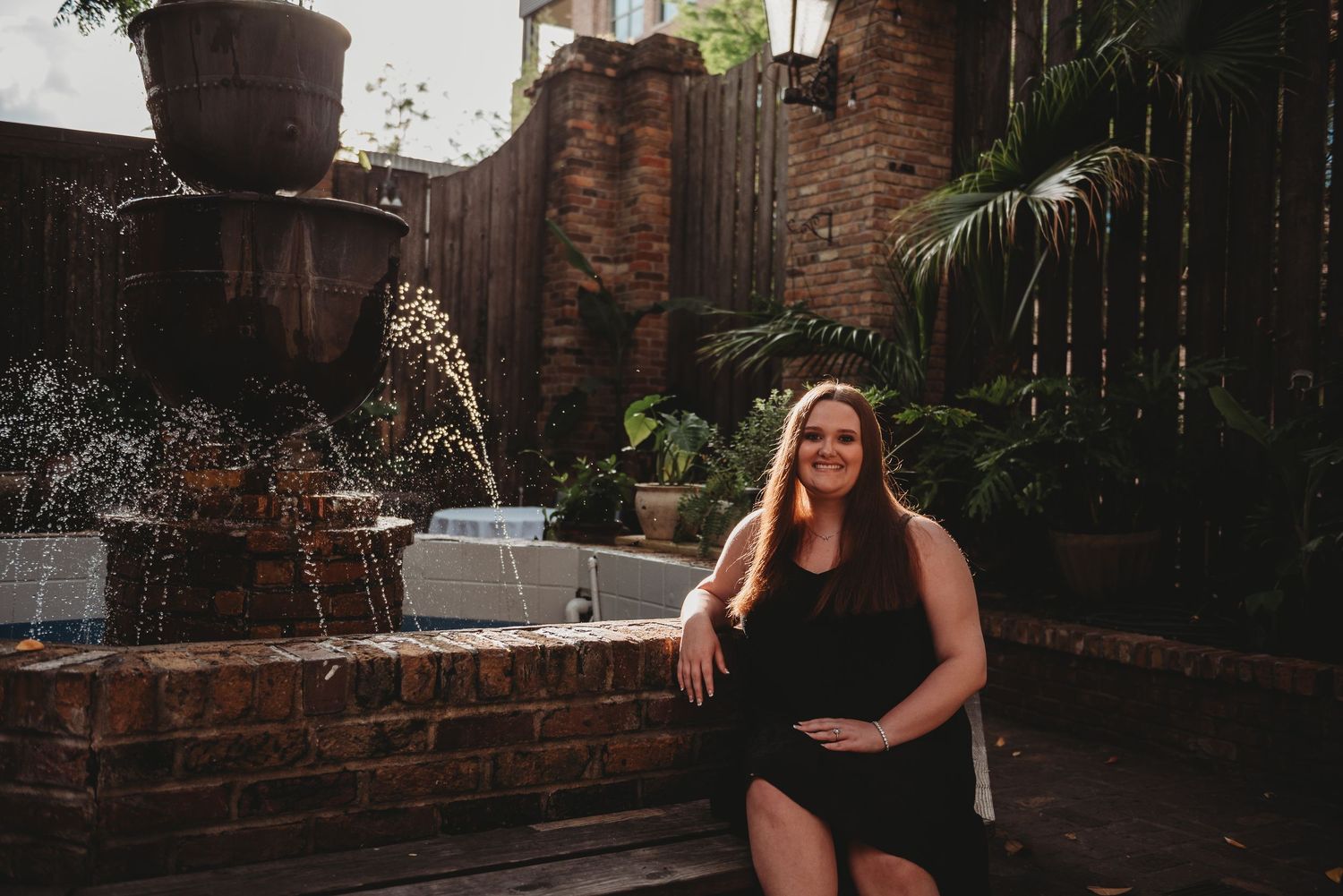 A smiling woman in a black dress sits beside a brick fountain in a lush courtyard.