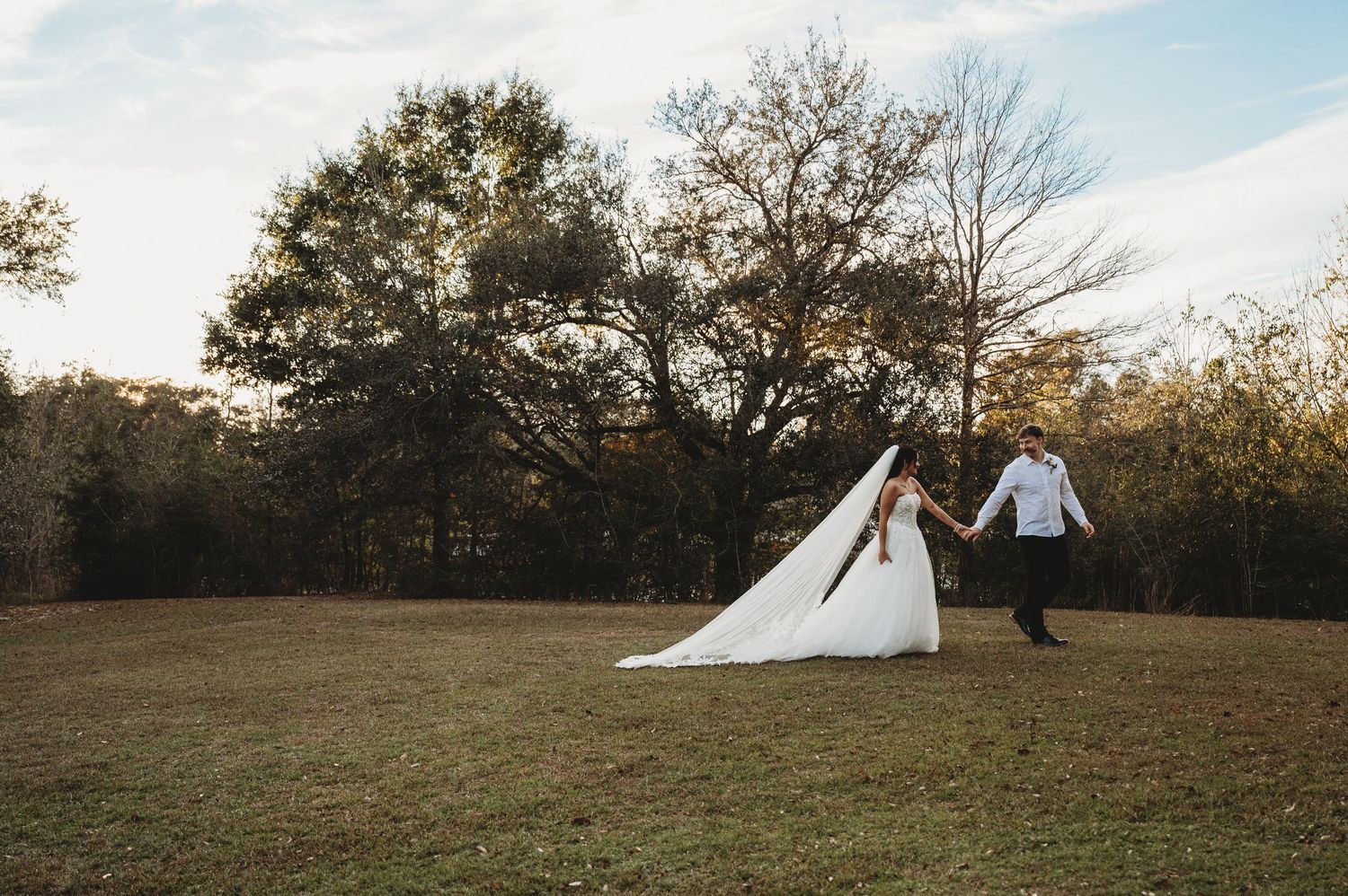 Bride and groom holding hands walking across a sunlit green field with autumn trees in the background.