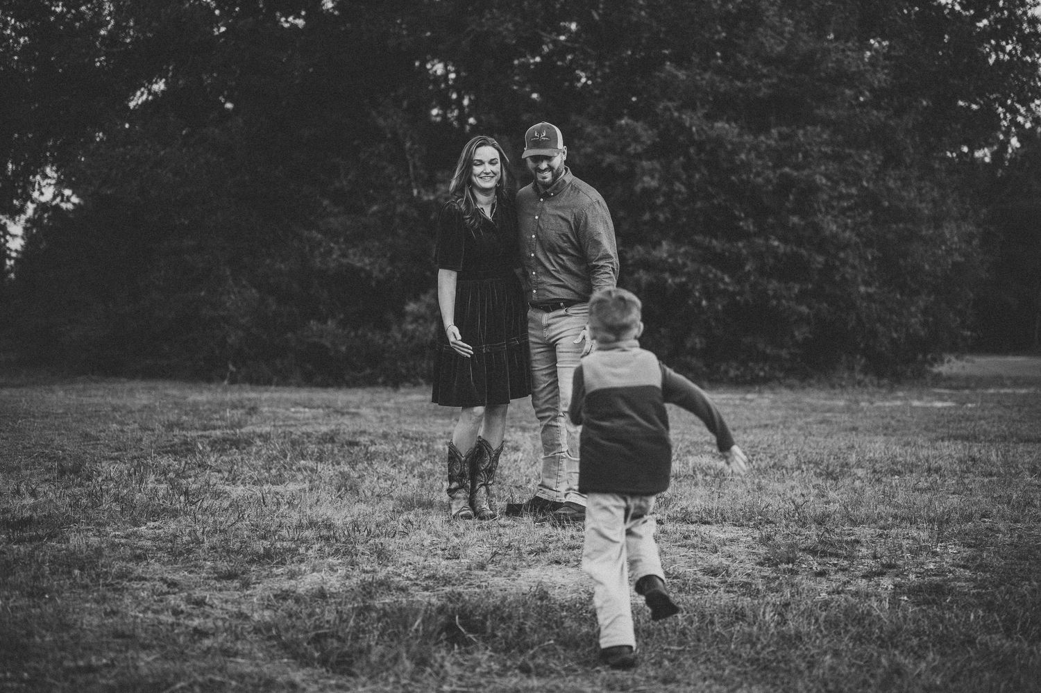 Black and white family photo of couple standing in grassy field as young child runs toward them.