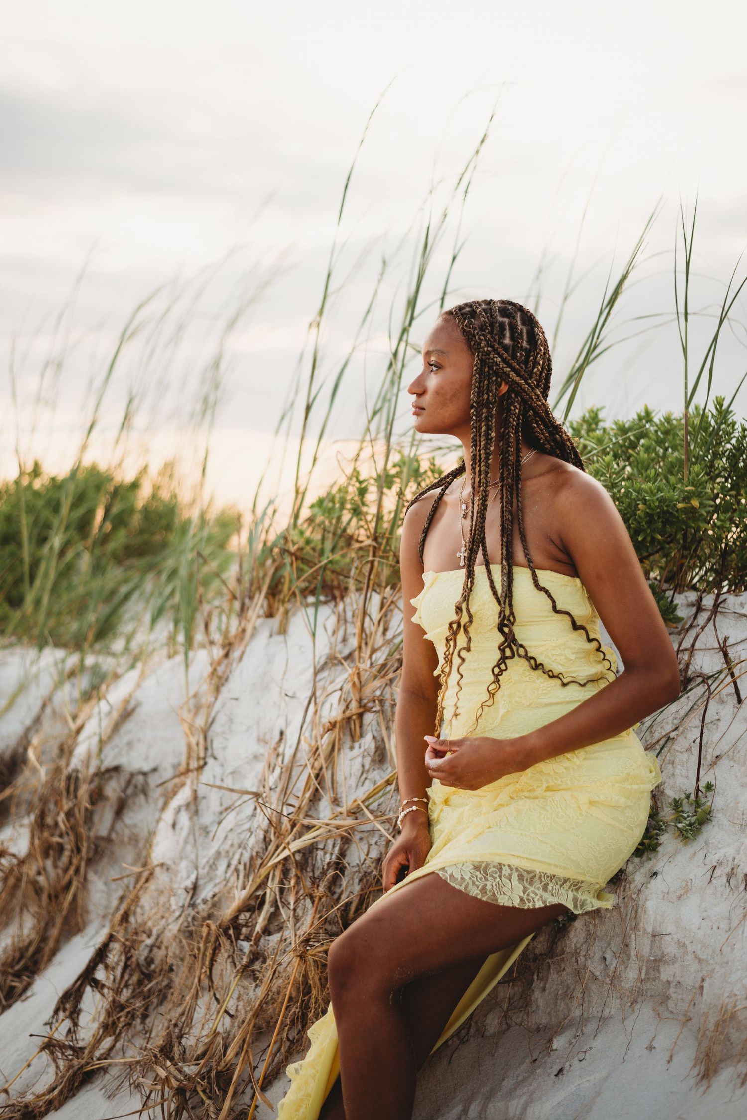 Young woman with braids in a yellow strapless dress sitting on sand dunes at sunset.