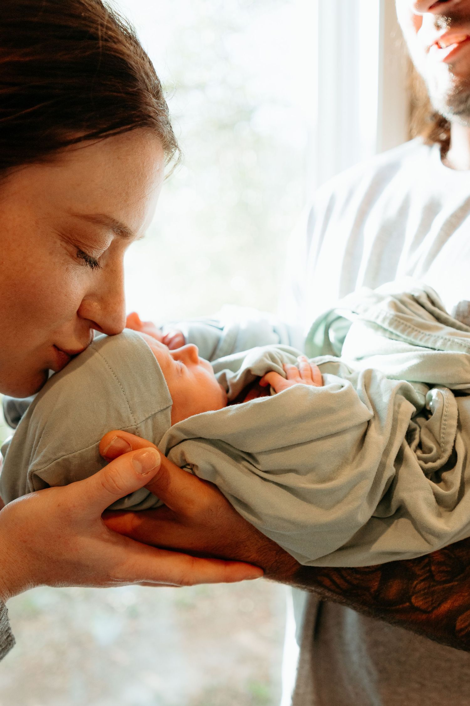 A new mother gently kisses her sleeping newborn baby wrapped in a soft green blanket.