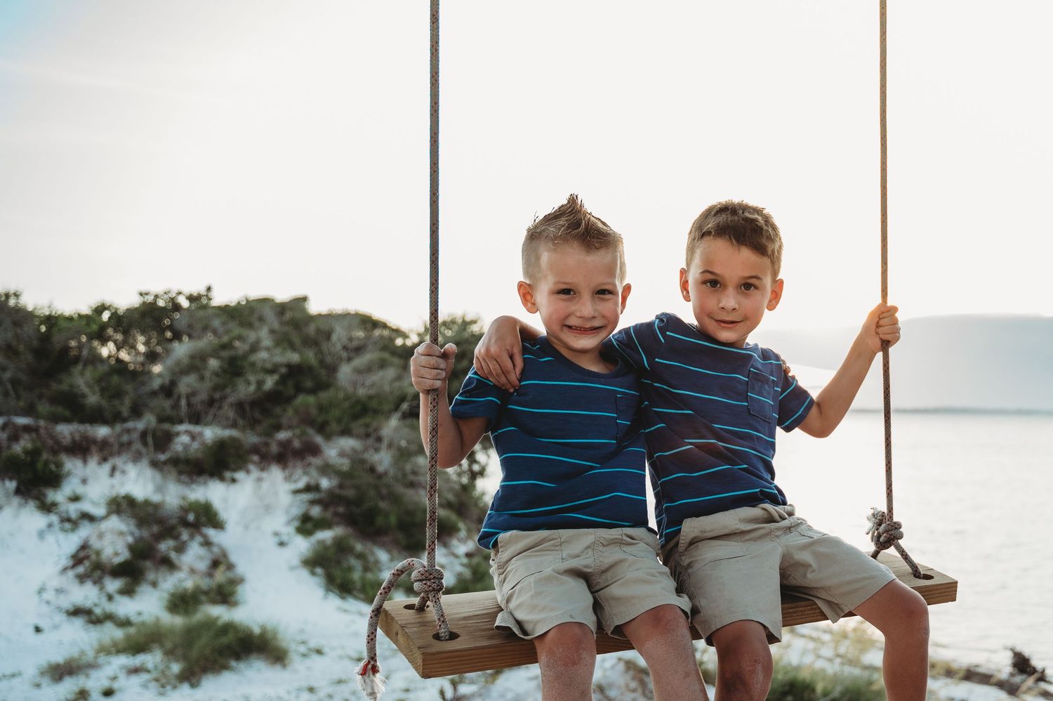 Two young boys smile together on a wooden swing at a sandy beach.