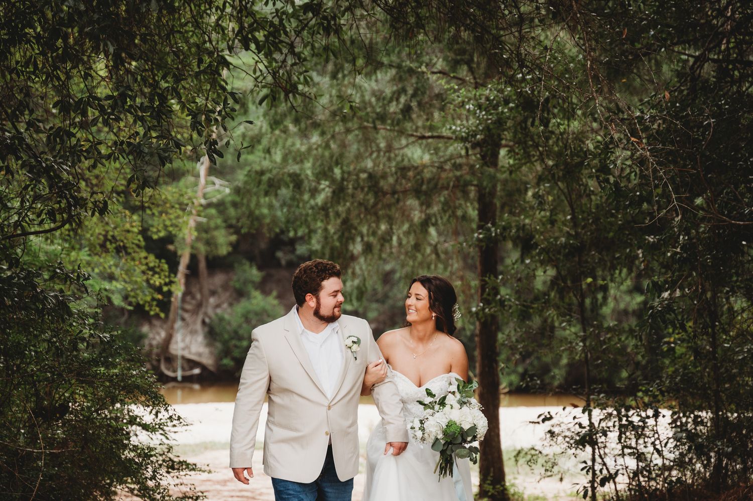 Bride and groom walking hand in hand through a lush green tree-lined path on wedding day.