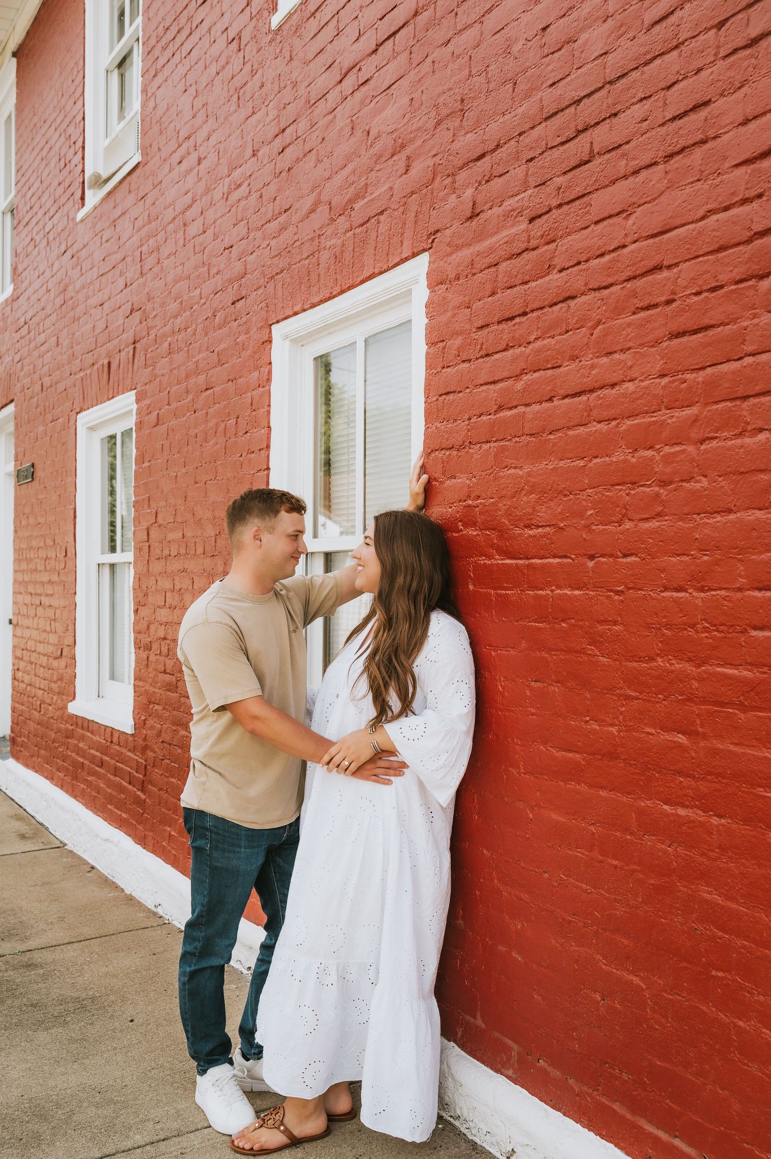 Couple sharing a romantic moment against a red brick wall, man in tan shirt and jeans, woman in white dress.