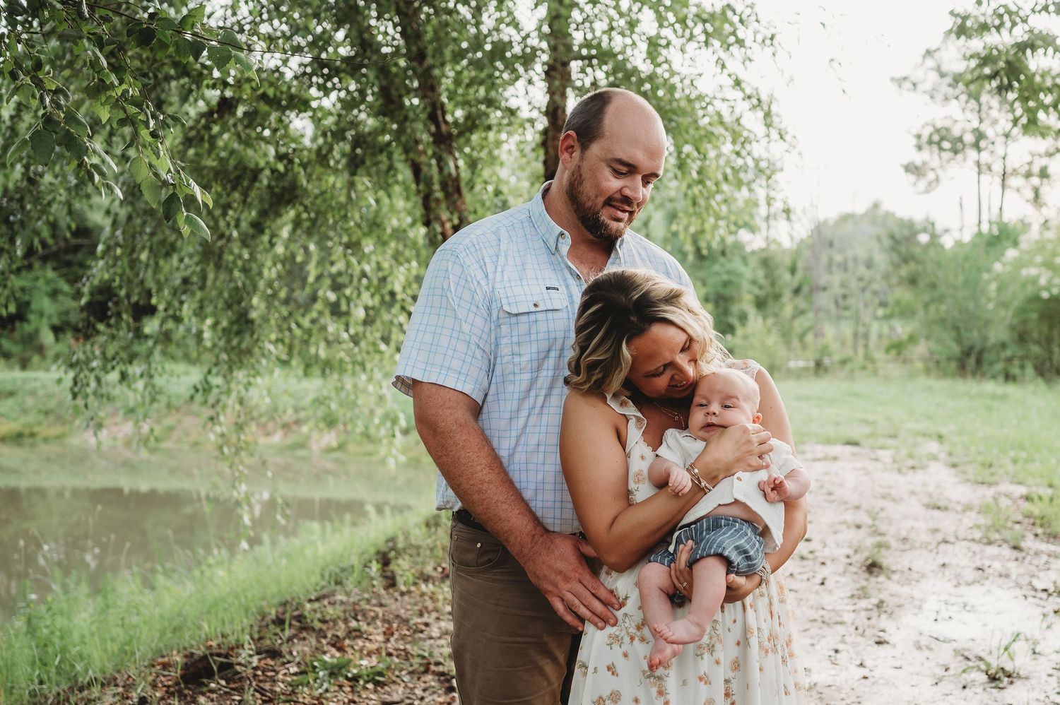 A happy family of three poses outdoors near a pond, with parents smiling and holding their baby girl.