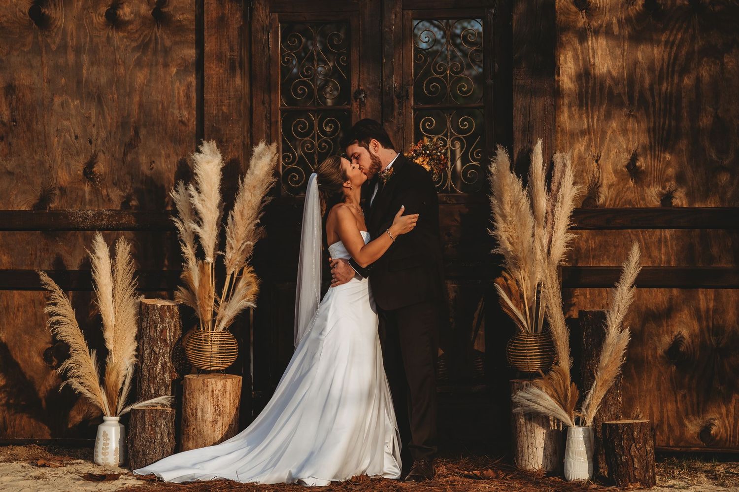 Bride and groom sharing a romantic kiss in front of rustic wooden doors surrounded by pampas grass arrangements.