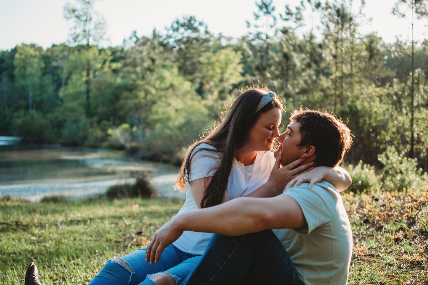 Couple sharing a tender kiss while sitting together on grass near a peaceful lake surrounded by trees.