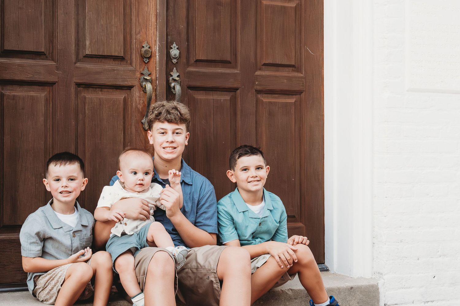 Teenage boy holding a baby flanked by two younger brothers on brick steps before wooden doors.