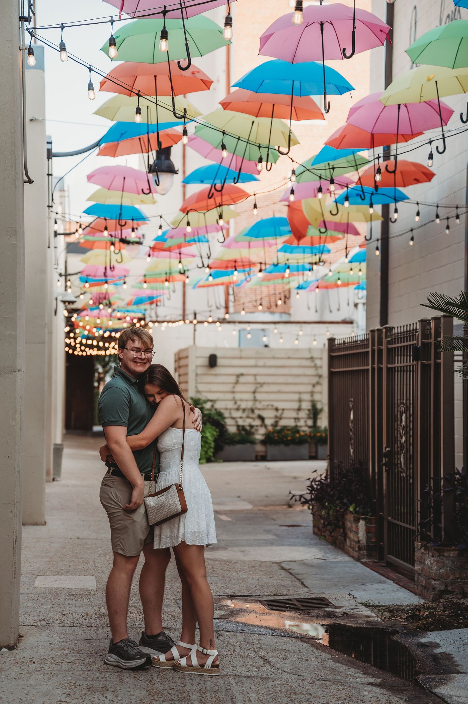 Couple embracing under colorful umbrella installation in a charming alleyway decorated with string lights.