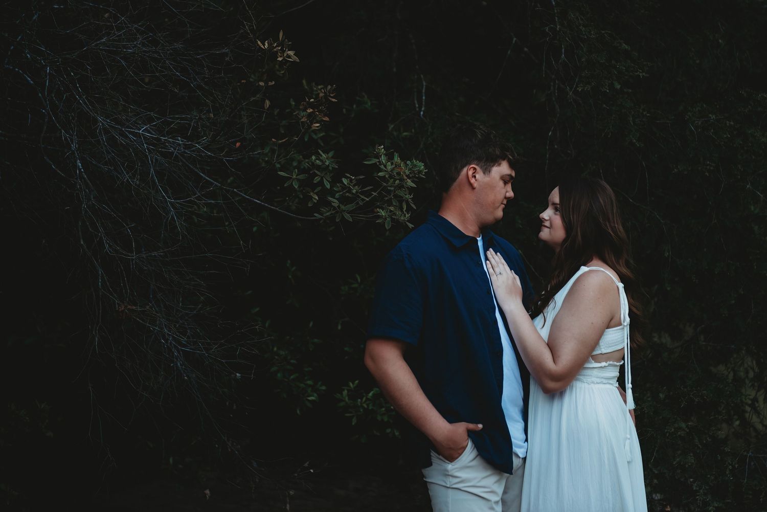 Couple gazing into each other's eyes in a dark, moody outdoor setting with lush foliage behind them.
