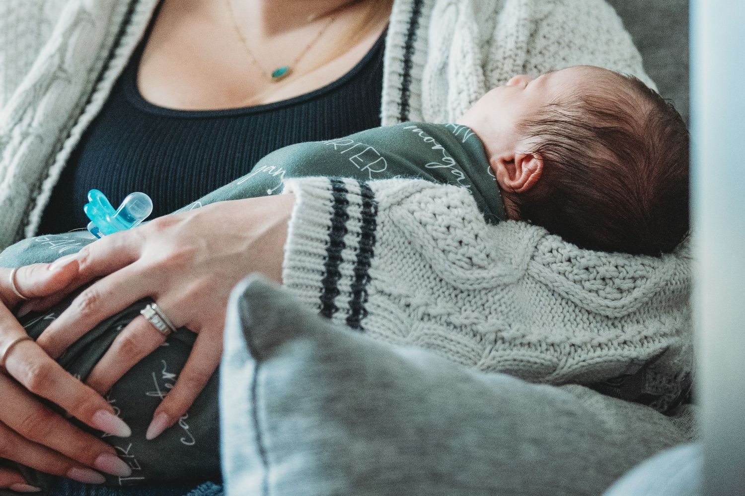 Mother cradling sleeping newborn baby dressed in gray knit outfit.