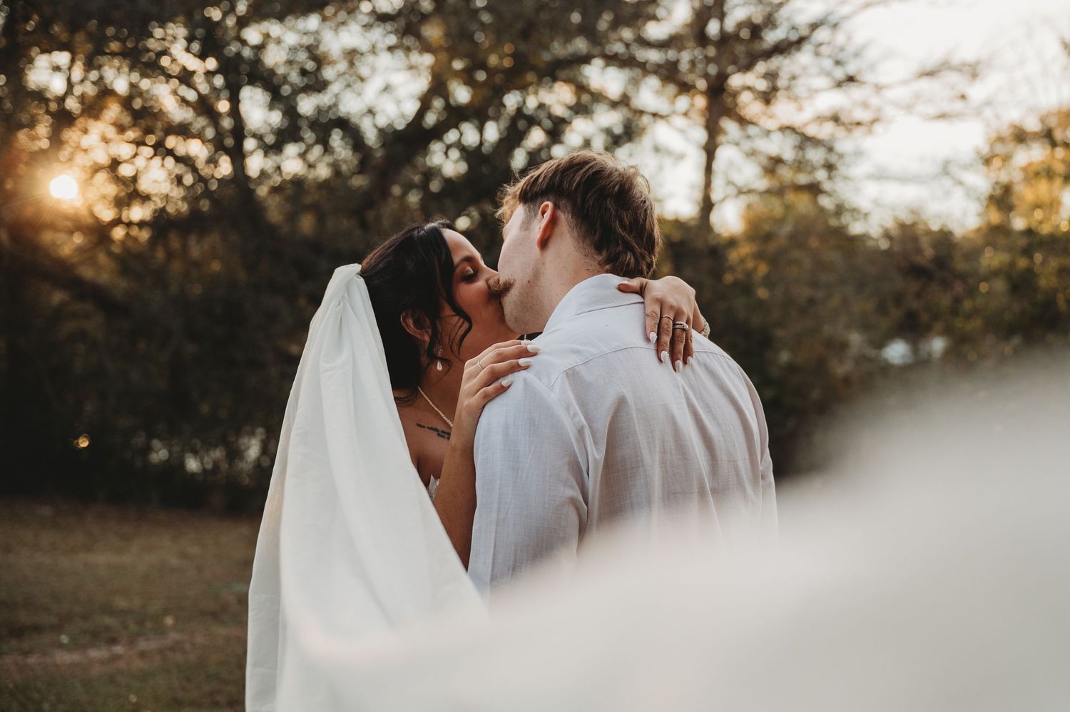 A bride and groom share a romantic kiss outdoors at golden hour, with the brides veil flowing in the breeze.