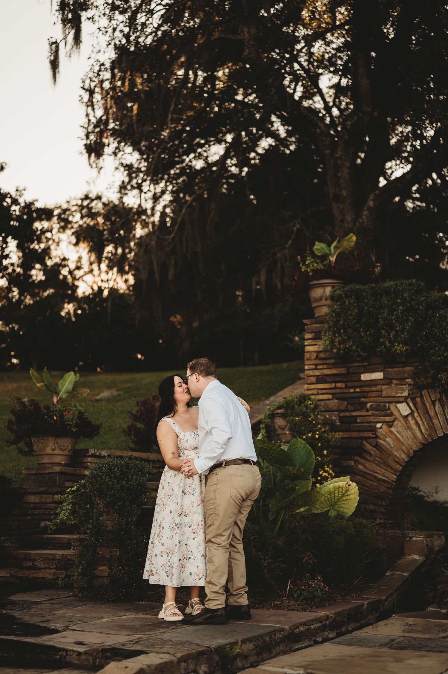 Couple sharing a kiss in a lush garden at sunset with stone archway.
