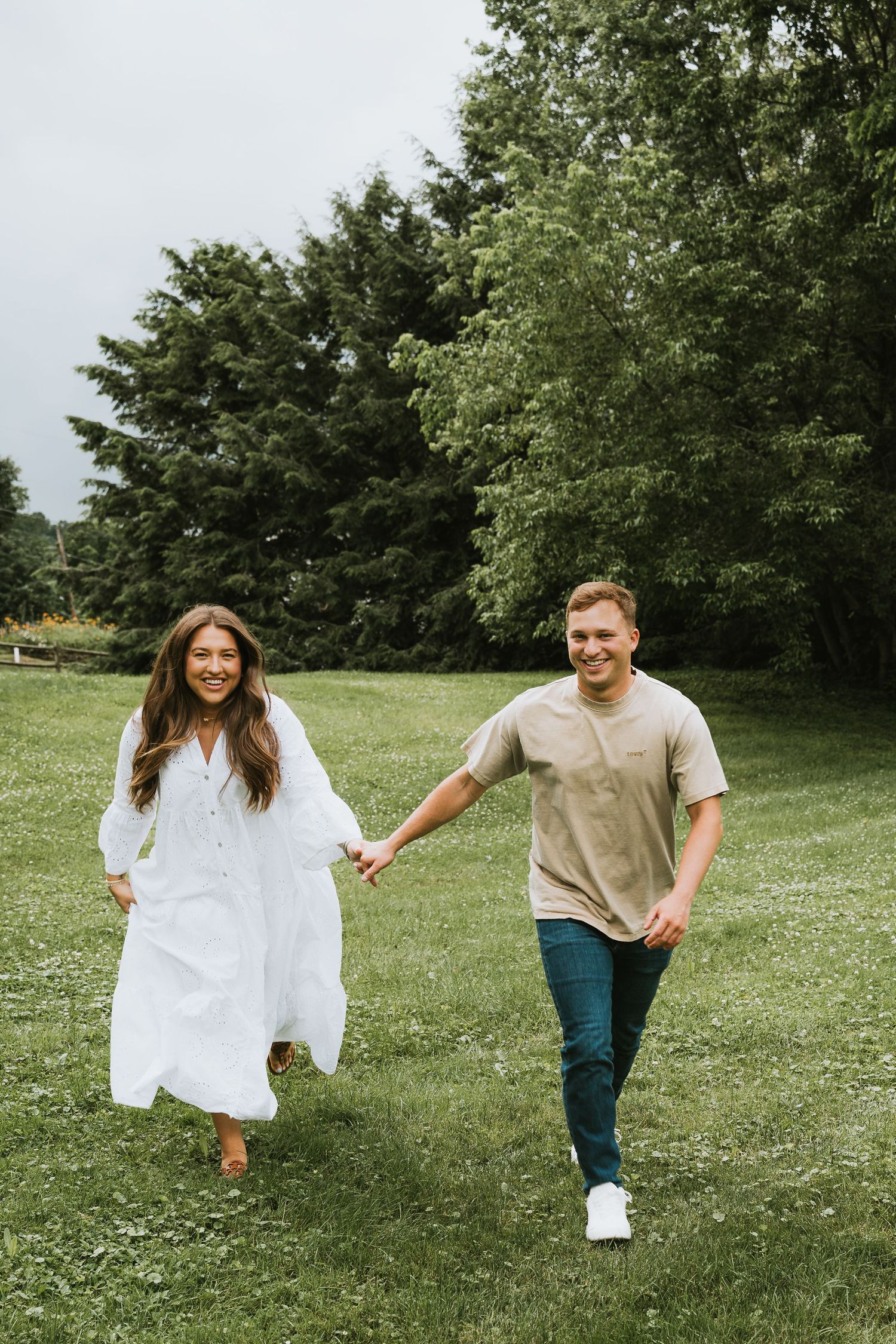 A smiling couple holds hands while walking through a lush green park, woman in white dress, man in beige shirt.
