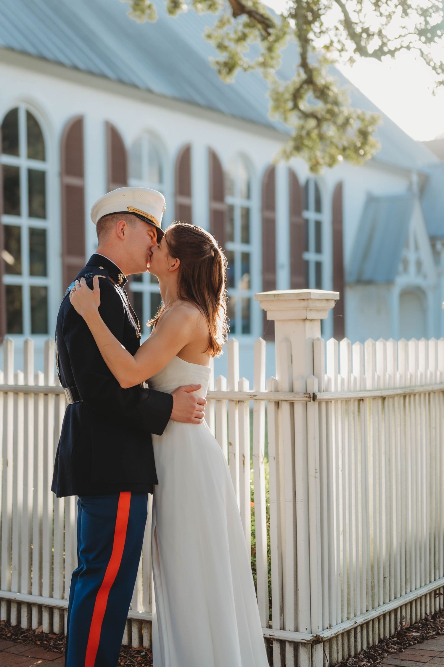 Marine in dress uniform kisses bride in white gown by white picket fence near historic building.