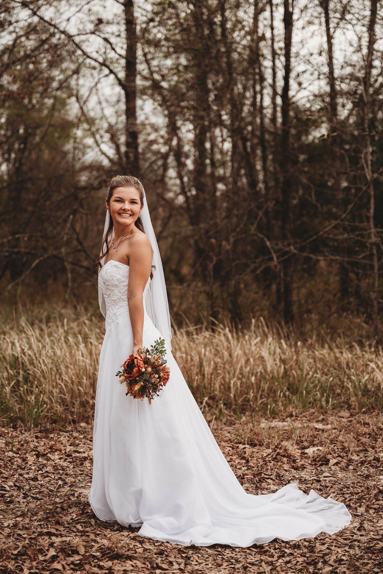 Smiling bride in white strapless gown and veil holds autumn bouquet in a fall forest setting.