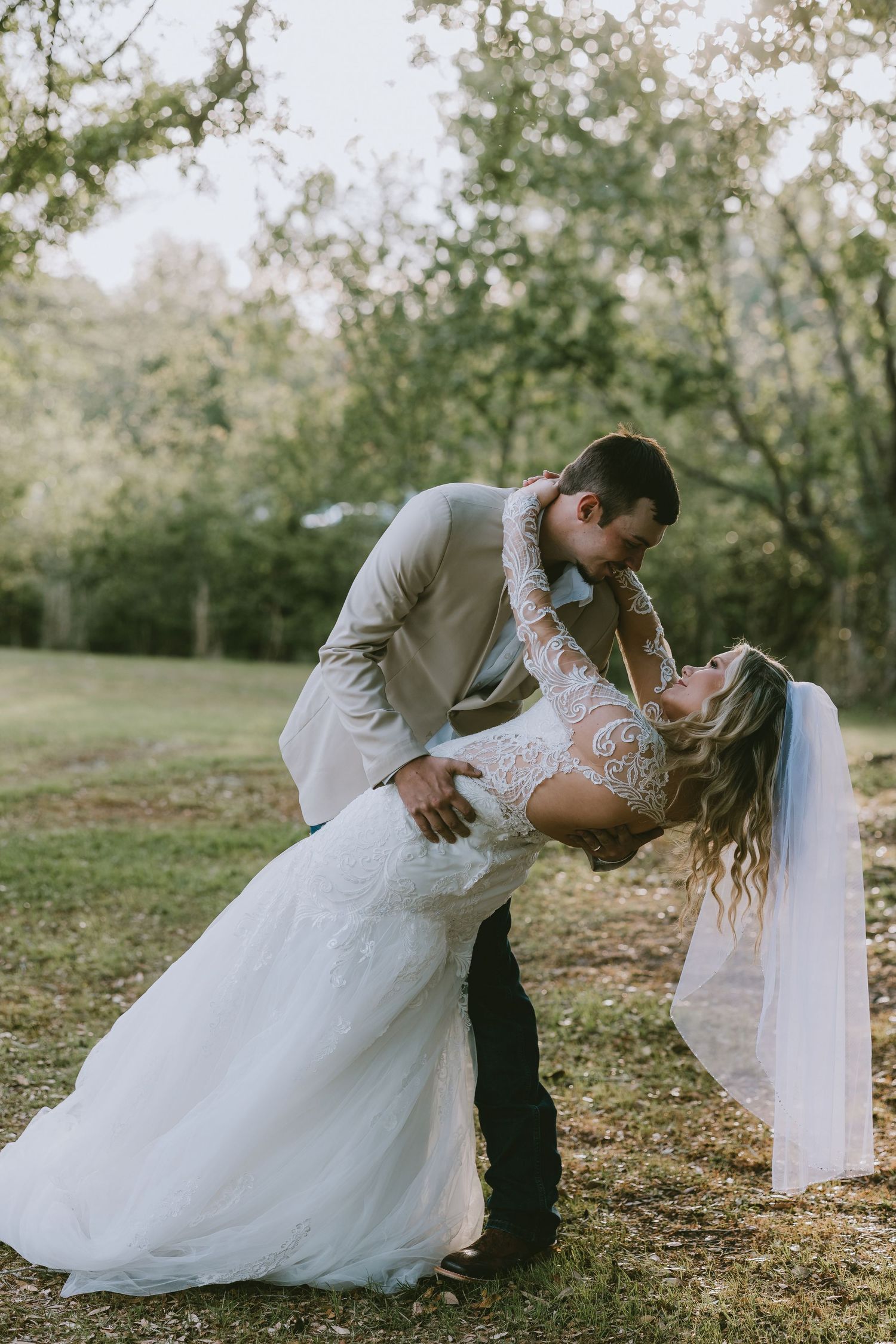 Groom dipping bride for a romantic kiss outdoors, bride in lace long-sleeve wedding gown with veil.