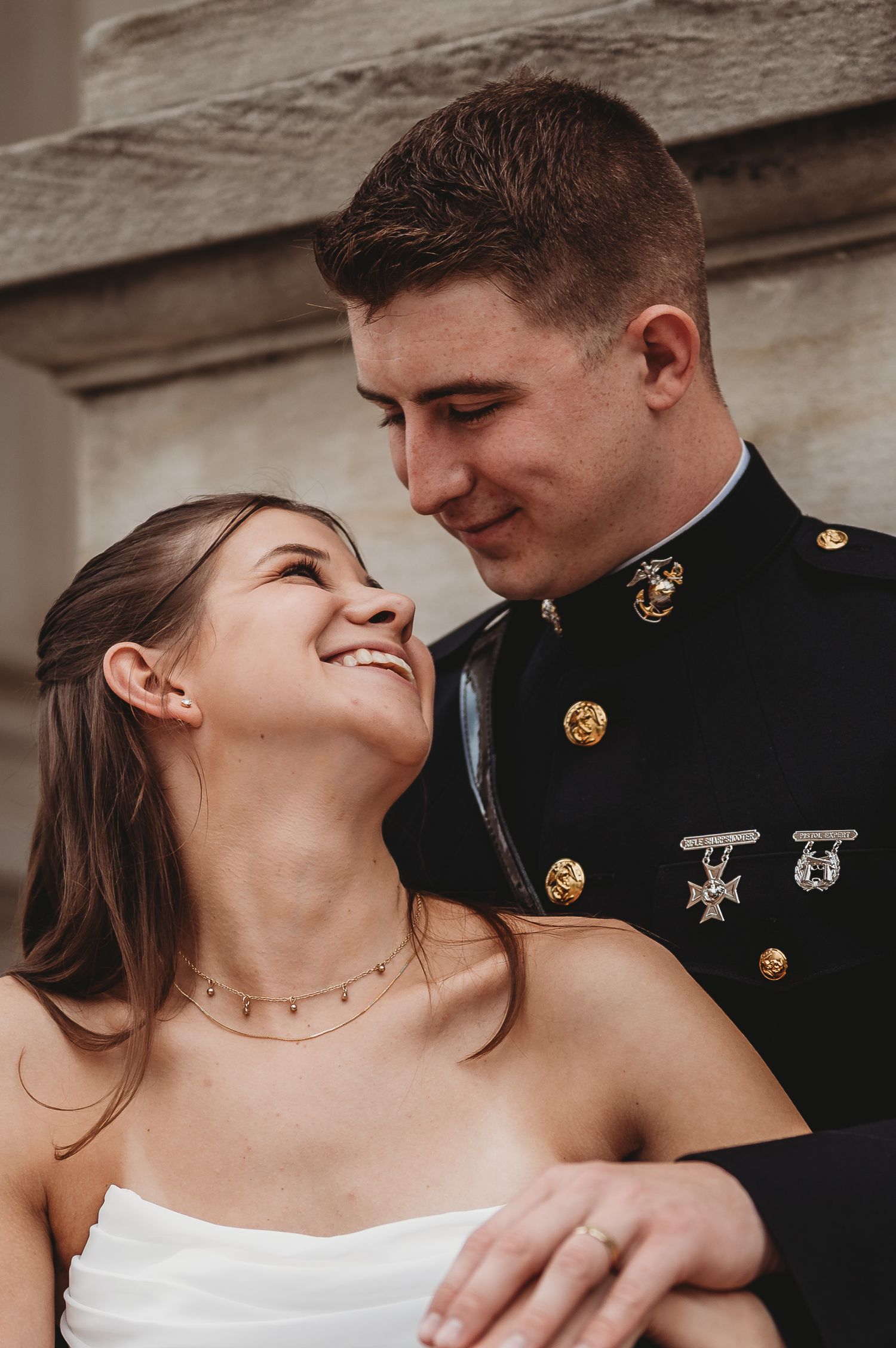A smiling bride in white looks up at a Marine in dress blues, sharing a romantic moment together.