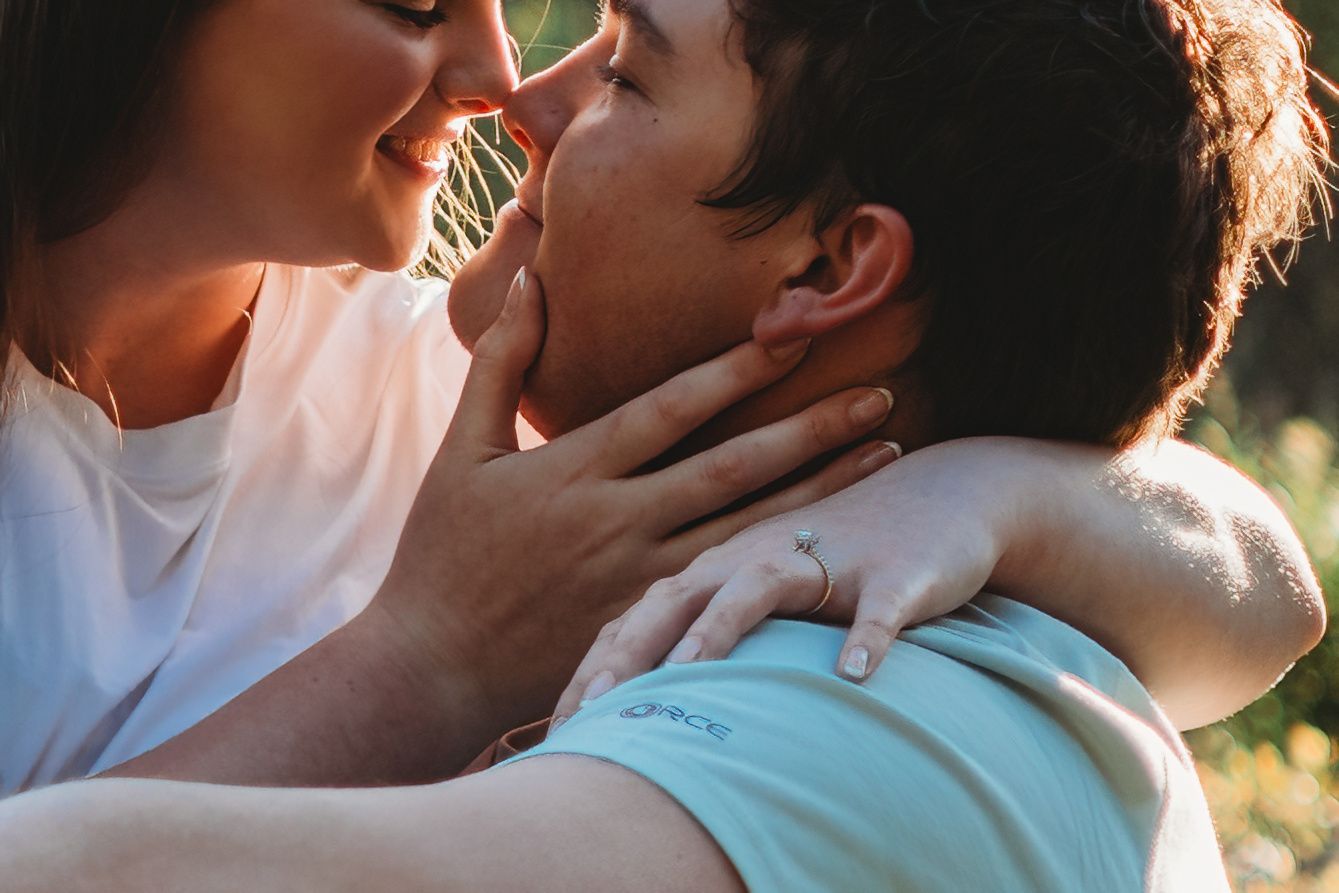 Couple sharing a romantic almost-kiss outdoors, woman wearing engagement ring embracing man in warm golden light.