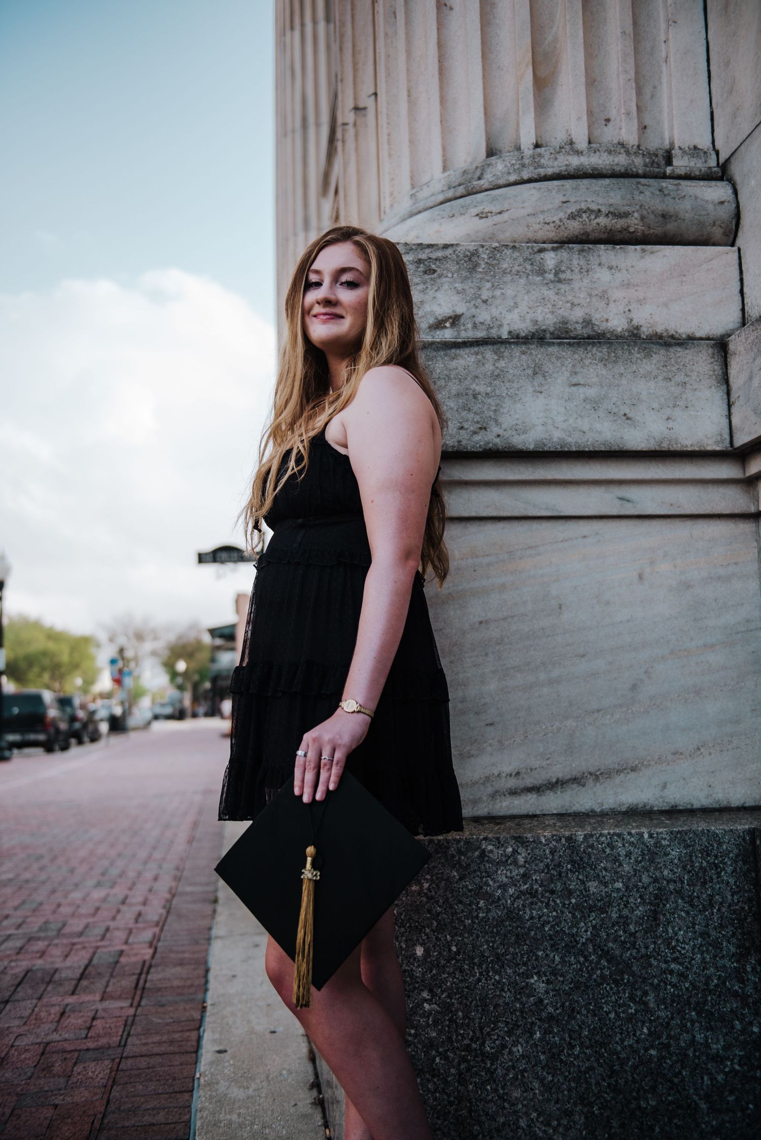 Young woman in black dress leans against stone column holding graduation cap on city sidewalk.