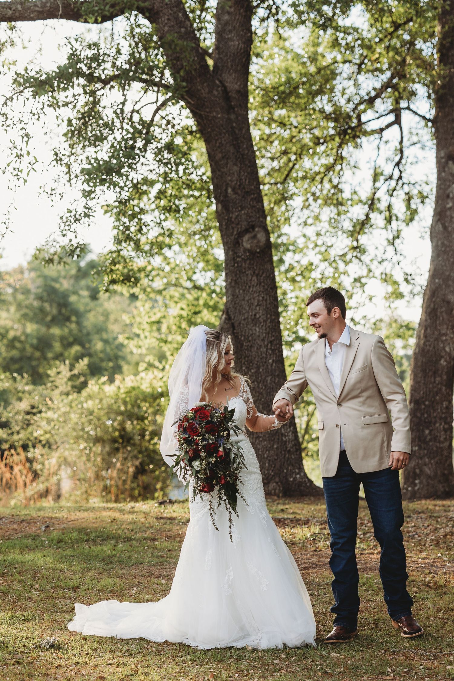 Bride and groom holding hands and smiling beneath a large oak tree during outdoor wedding.