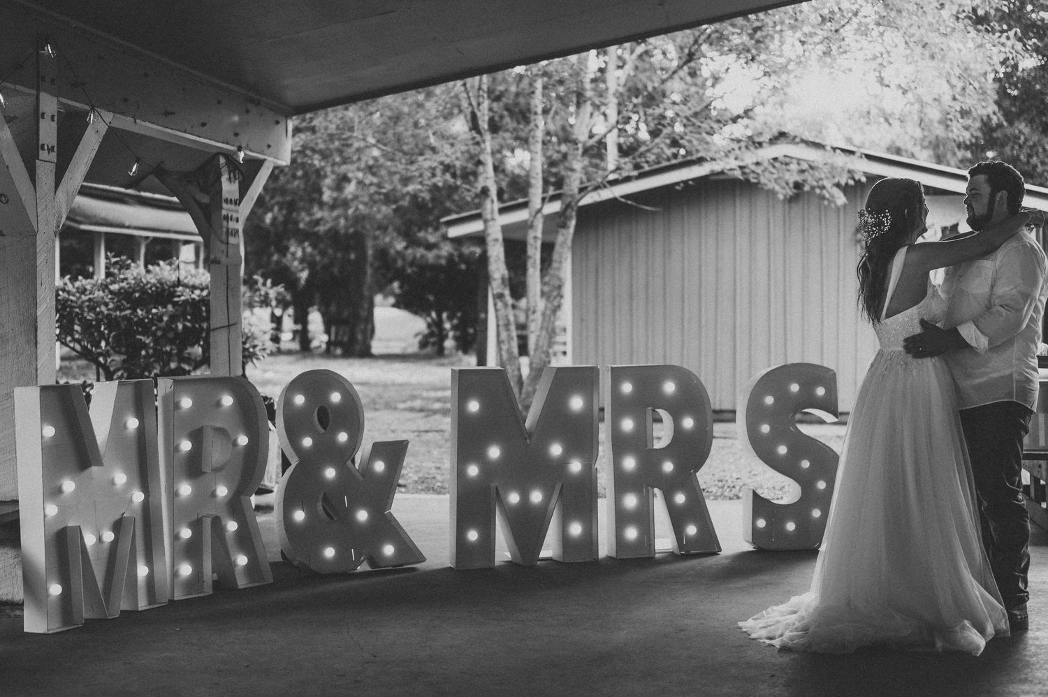 Black and white photo of newlyweds embracing behind large marquee MR & MRS letters with lights.