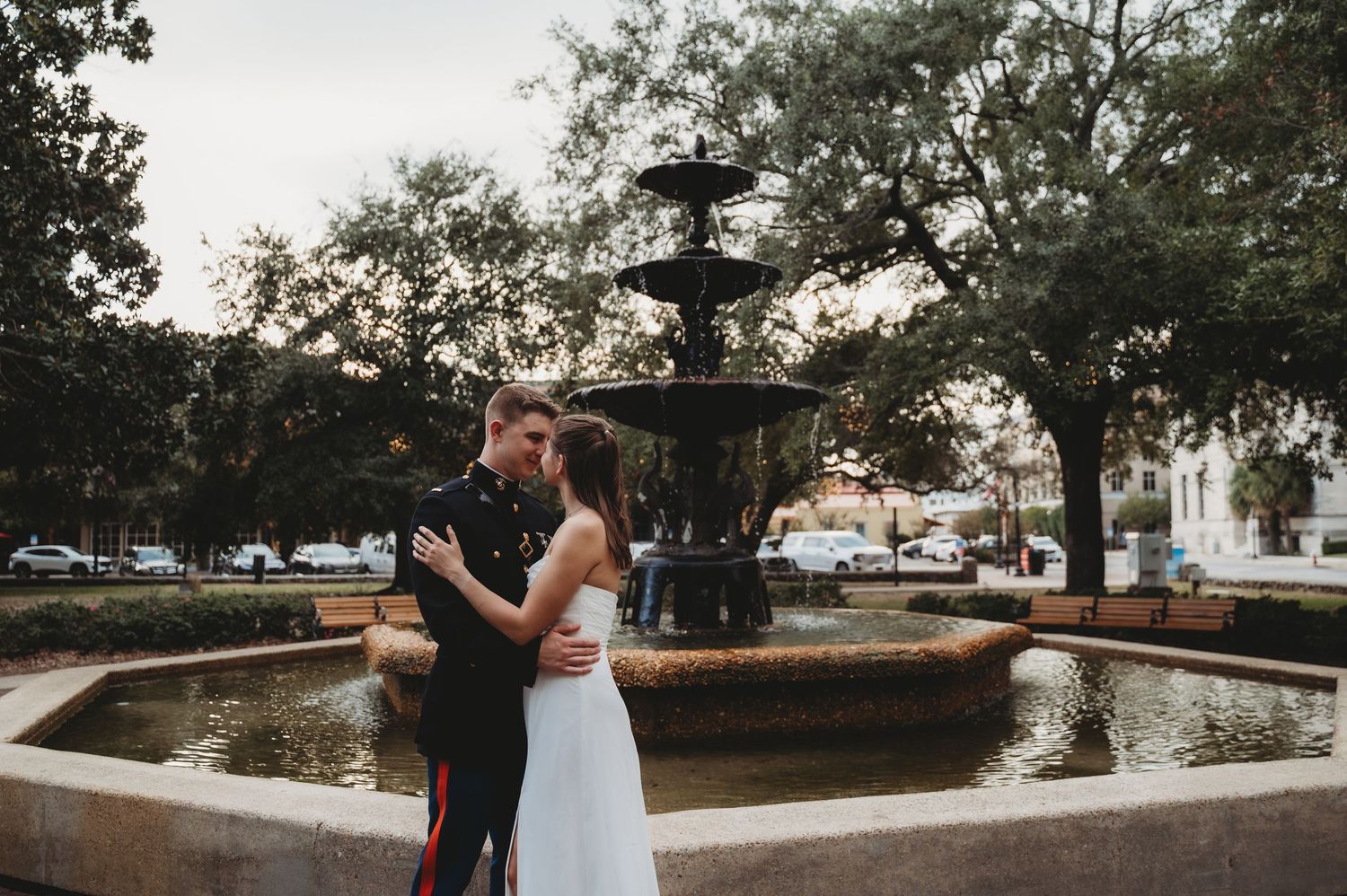 Newlyweds share romantic kiss by ornate fountain in scenic town square at dusk.