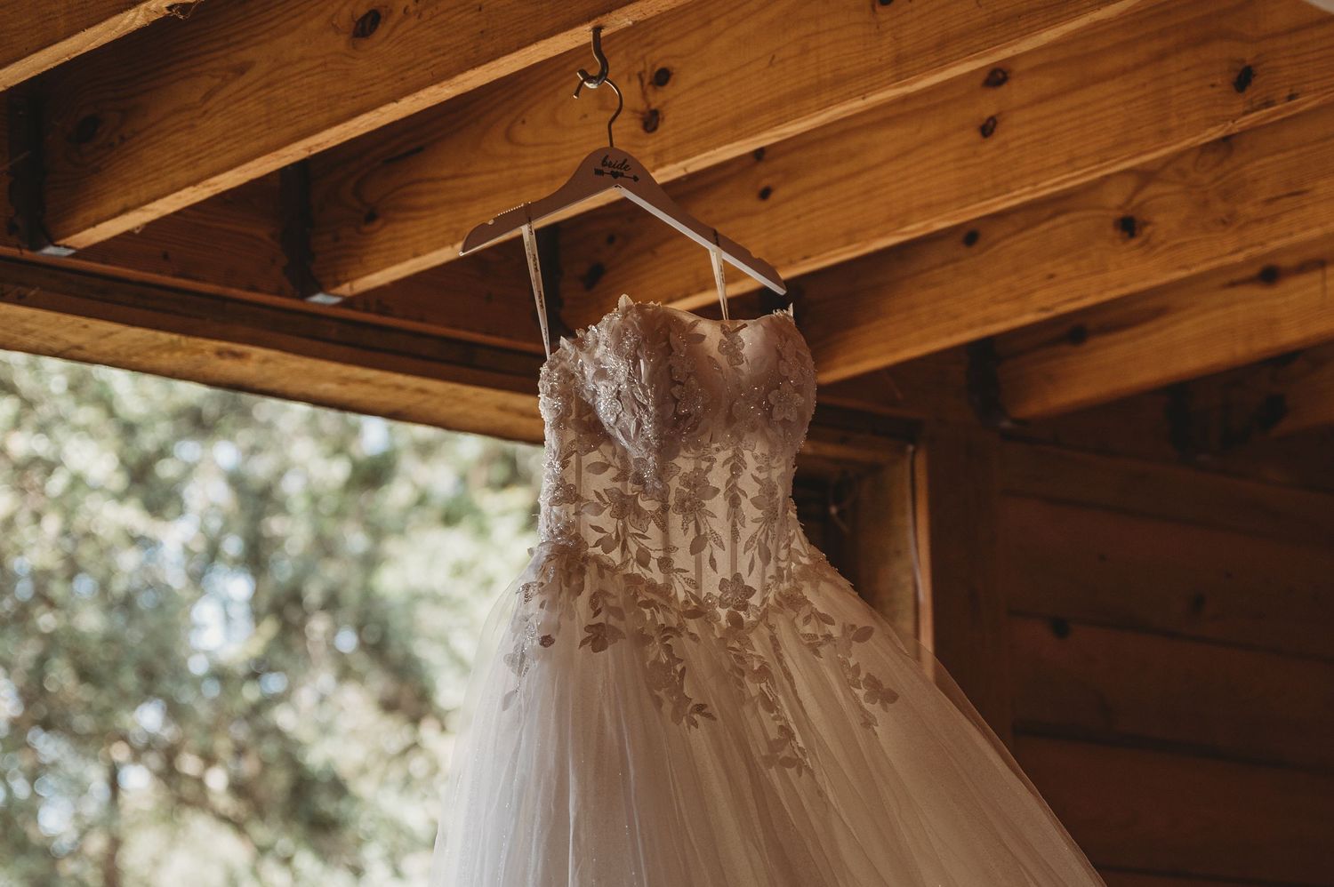A lace and tulle wedding dress hanging on a hanger from wooden cabin rafters with trees visible in background.