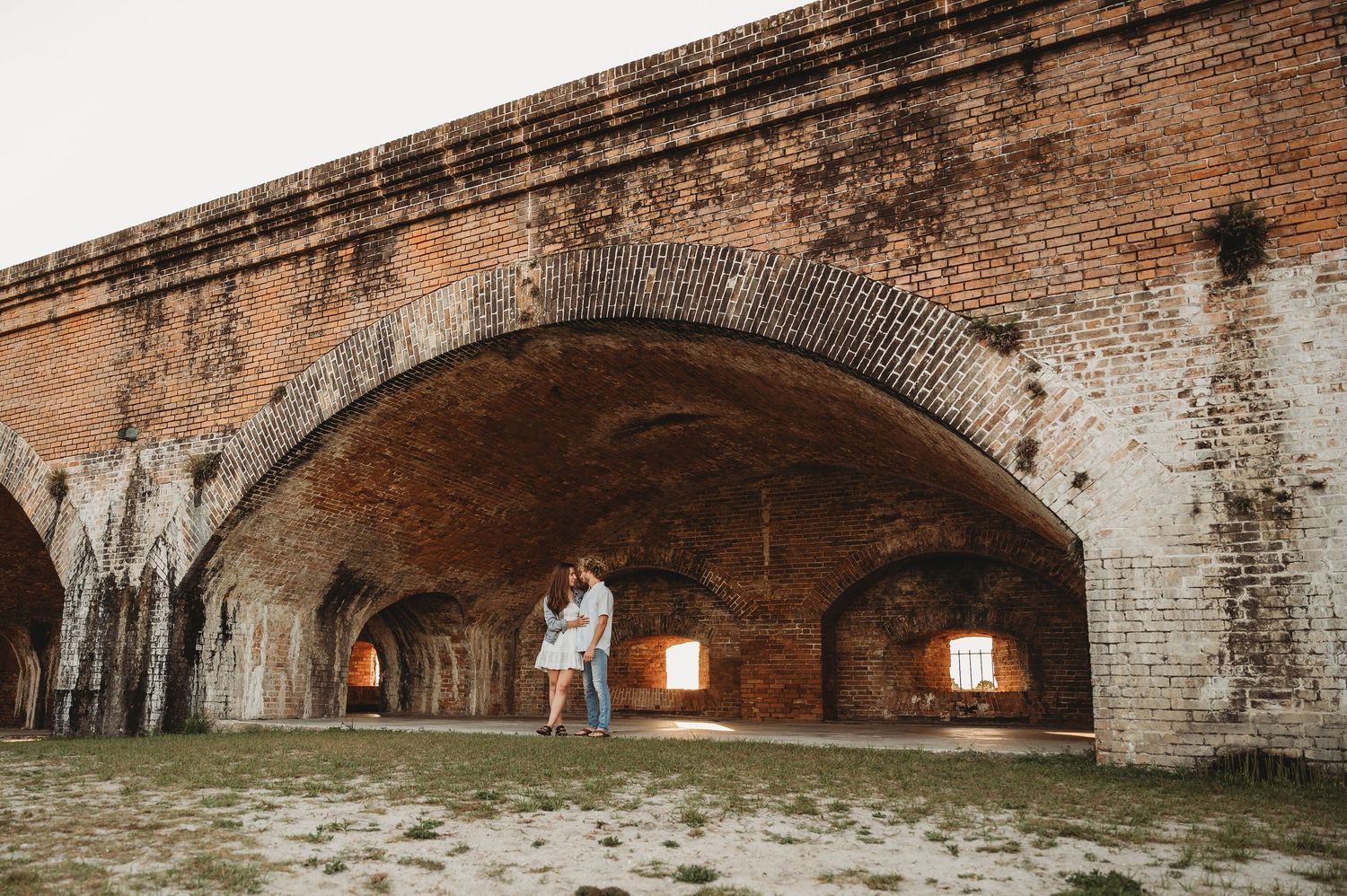 Romantic couple kissing under historic brick arched corridors in soft natural light.
