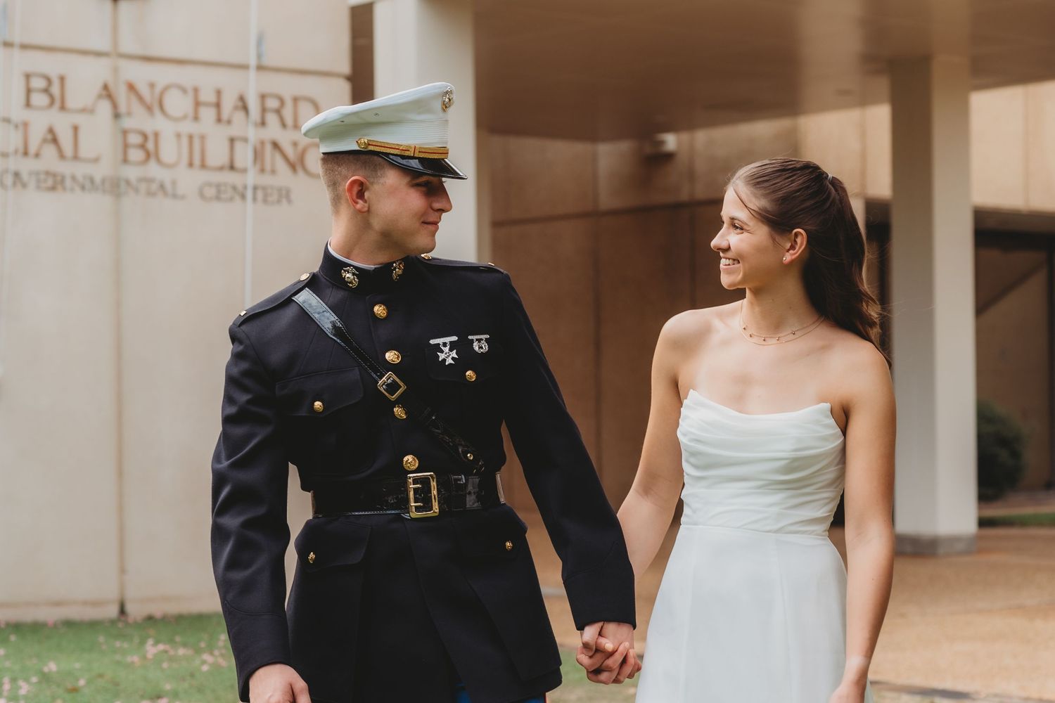 Marine in dress uniform holds hands with bride in white gown outside Blanchard Building on their wedding day.